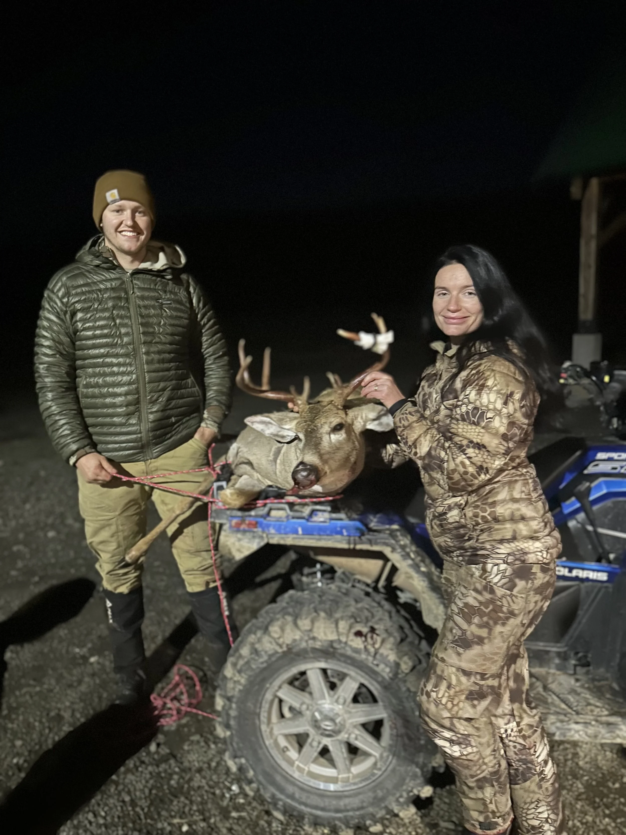 A man and woman outdoors at night with a deer on an ATV, the woman holding the deer's antlers.