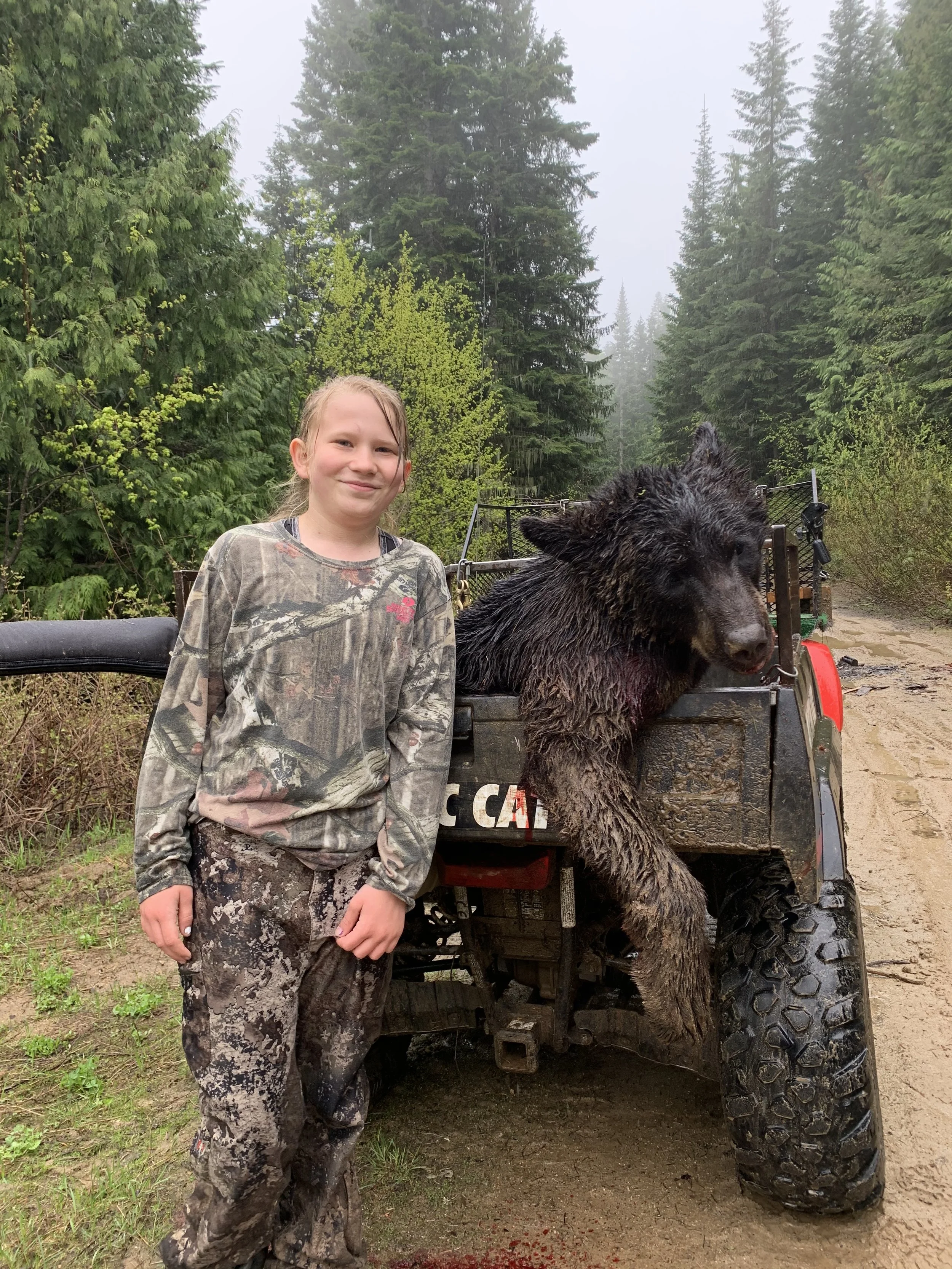 A young girl standing next to a bear carcass on the back of a vehicle in a forested area.