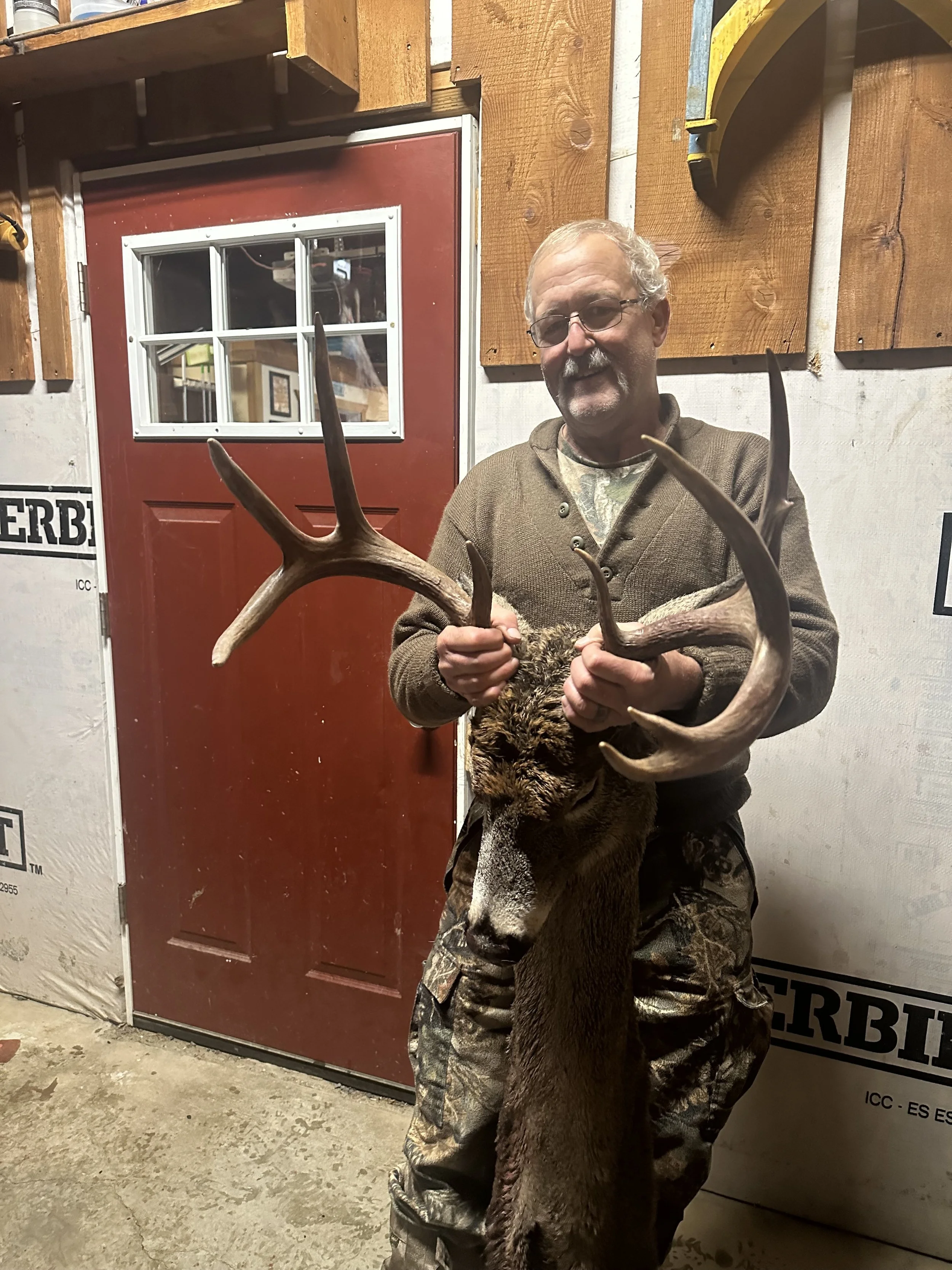 A man with glasses and a beard is holding a large deer head with antlers indoors, standing in front of a red door and a wall with wood paneling and insulation.
