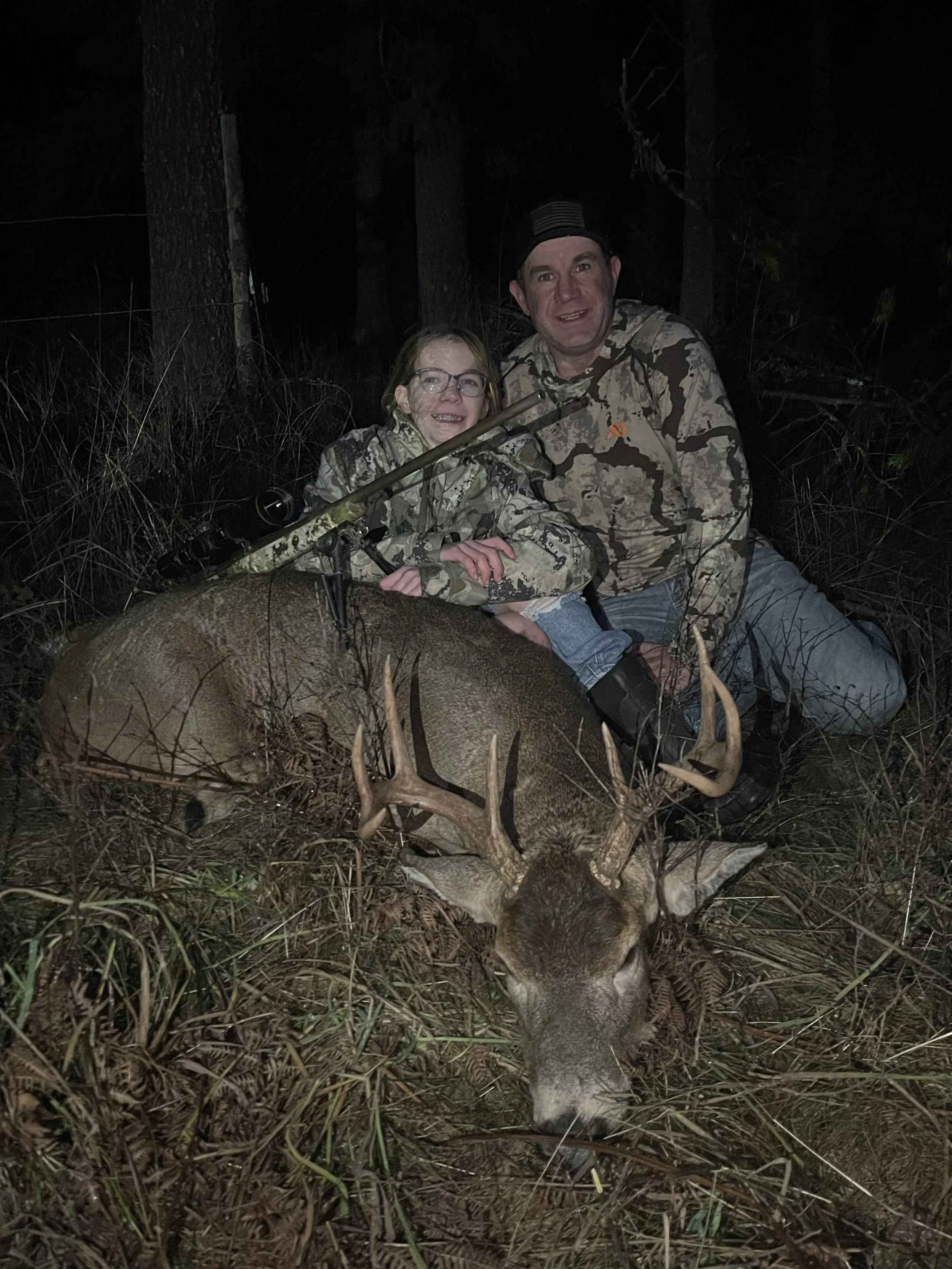 Two people, a man and a girl, sitting outdoors at night in a wooded area with a large, dead deer with antlers in front of them.