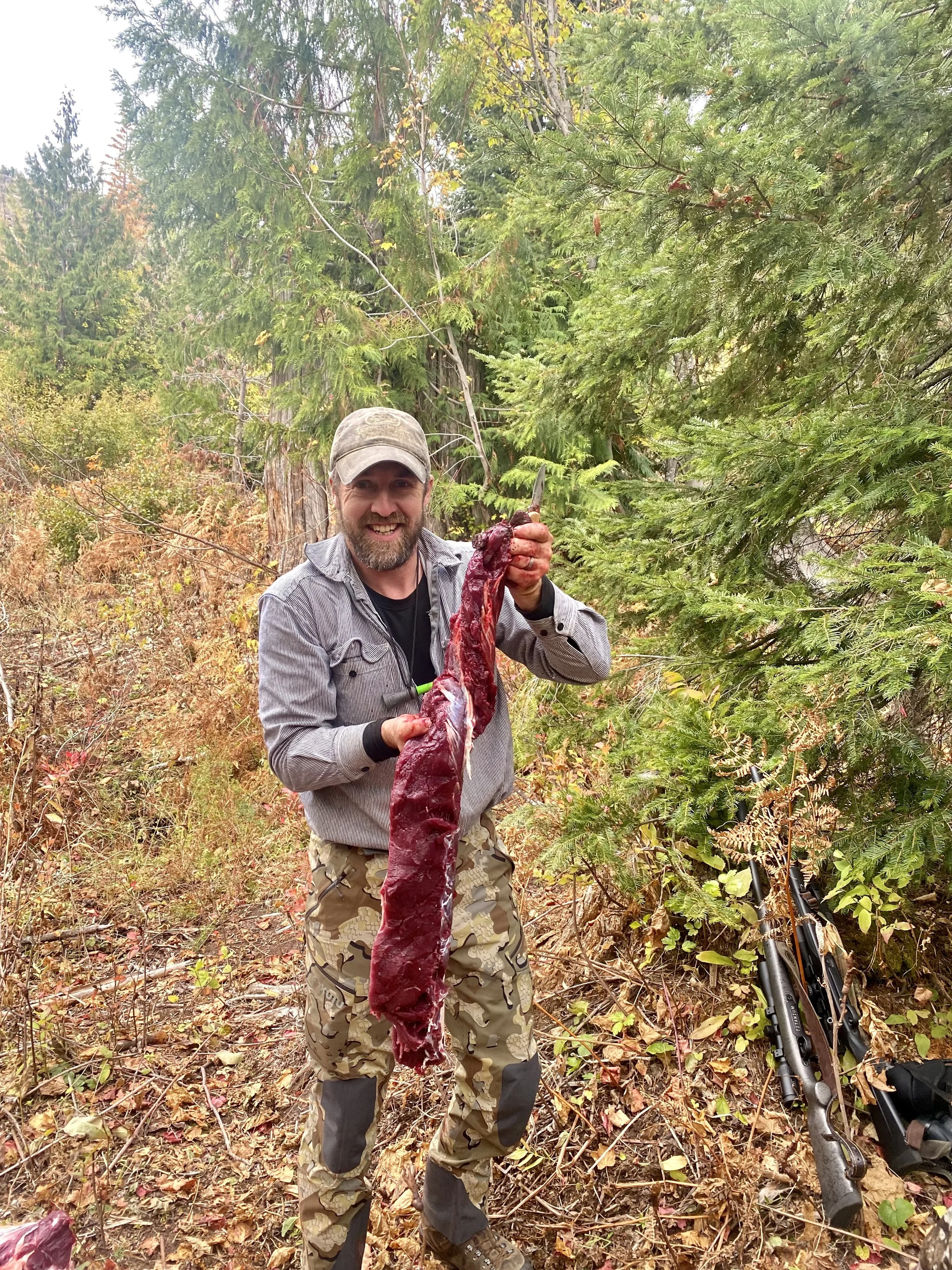 A man in outdoor clothing and camouflage pants standing in a forest, smiling, holding a long, raw piece of game meat with a knife in one hand. There are trees and fall foliage behind him, and a rifle on the ground nearby.