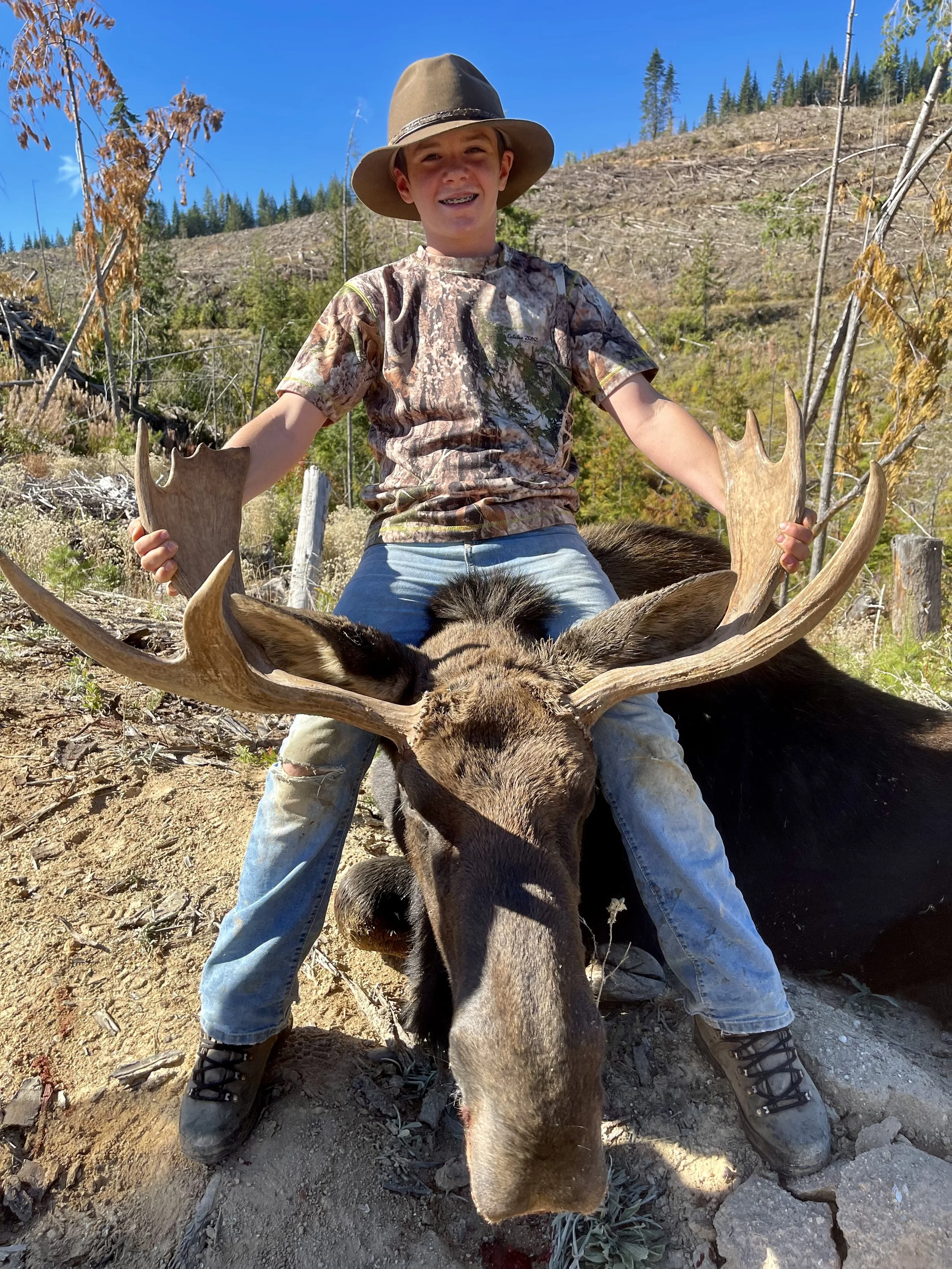 A boy wearing a wide-brimmed hat and camouflage shirt sits on a large moose carcass with antlers, smiling at the camera in a forested, mountainous area.