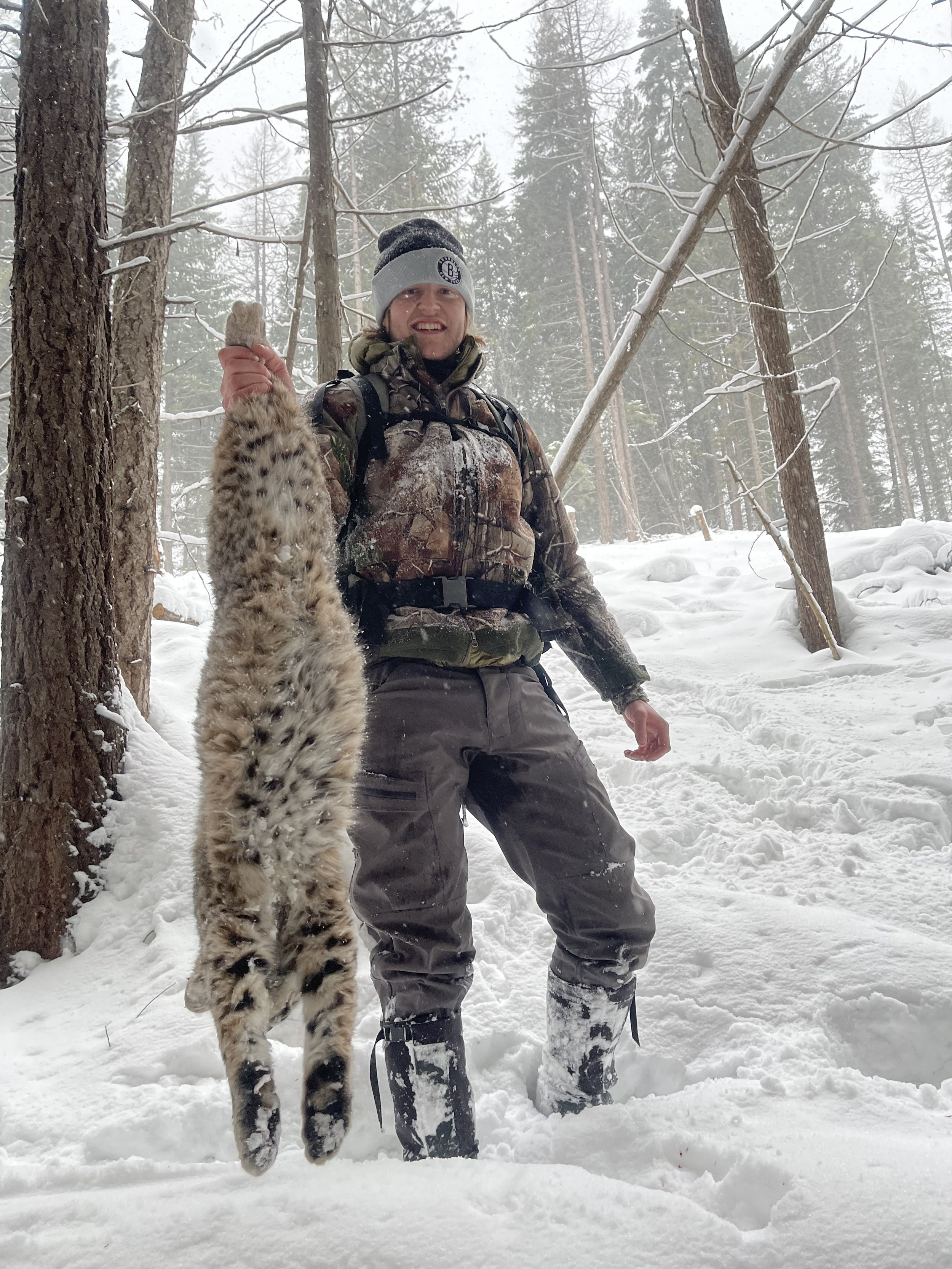 A man dressed in winter camouflage clothing and boots stands in a snowy forest, smiling and holding up a bobcat.