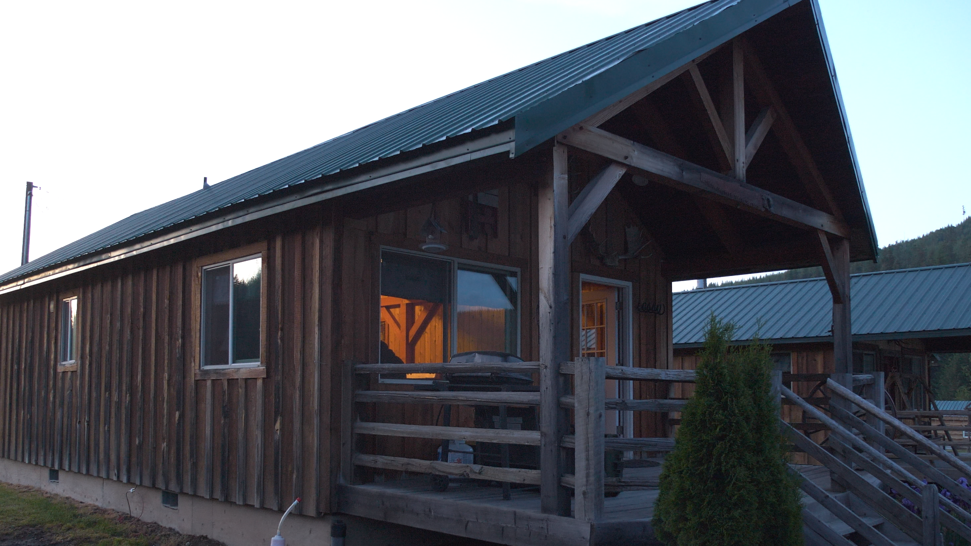 A rustic wooden cabin with a porch, illuminated from inside, with a grill on the porch, surrounded by trees and a hillside in the distance.