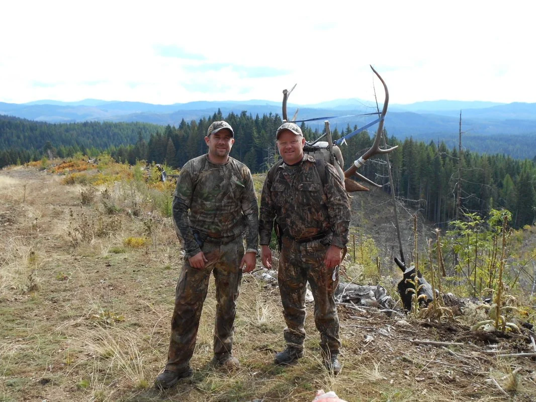 Two hunters in camouflage gear standing outdoors in a mountain landscape, next to a large set of antlers and a hunted animal.