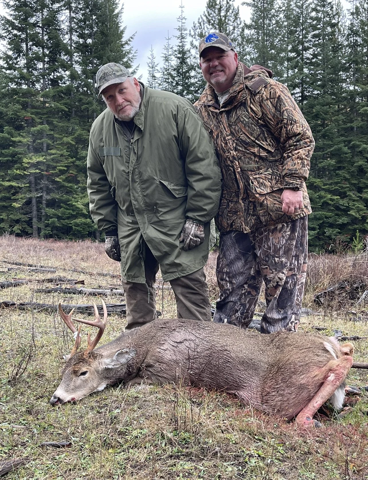 Two men in camouflage and outdoor gear standing behind a deer with large antlers, lying on the ground in a forested area.