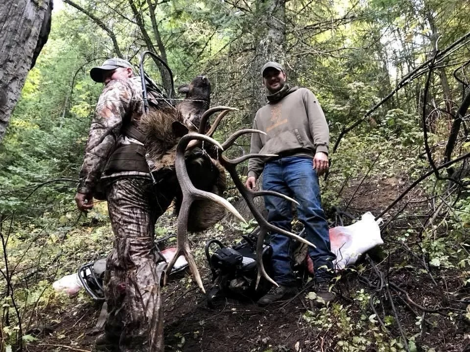 Two hunters in camouflage and outdoor gear stand in a forest, posing with a large elk they have hunted, with camouflage gear and backpacks around them.
