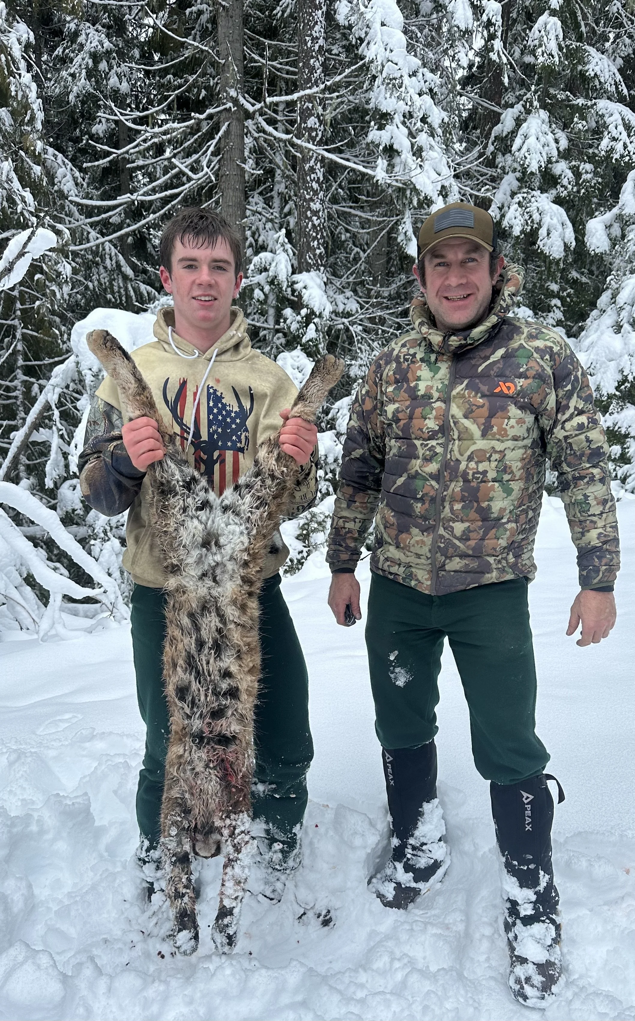 Two men standing in the snow in a forested area holding up a dead coyote, with snow-covered trees behind them.