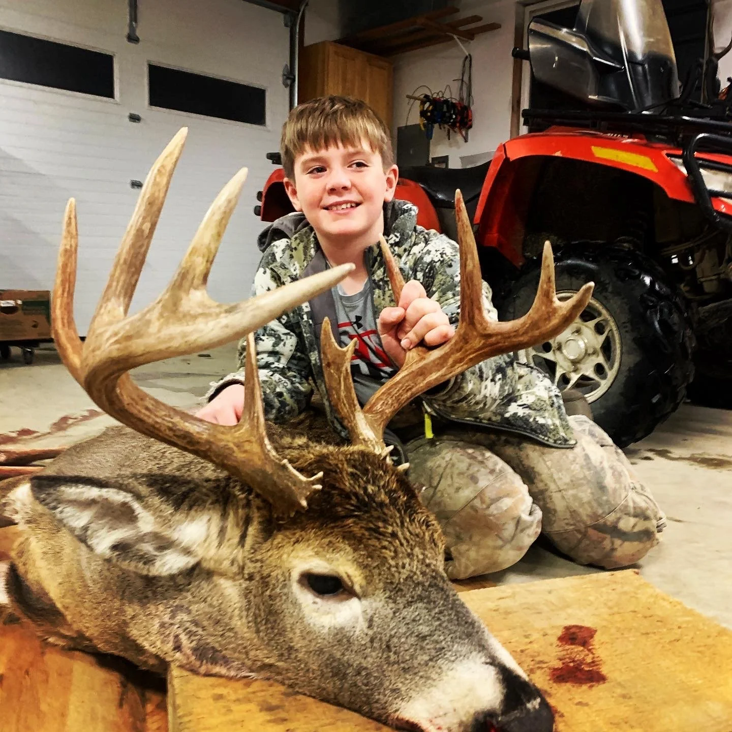 A boy in camouflage clothing kneeling behind a large deer with antlers, holding part of the antlers, inside a garage next to a red all-terrain vehicle.