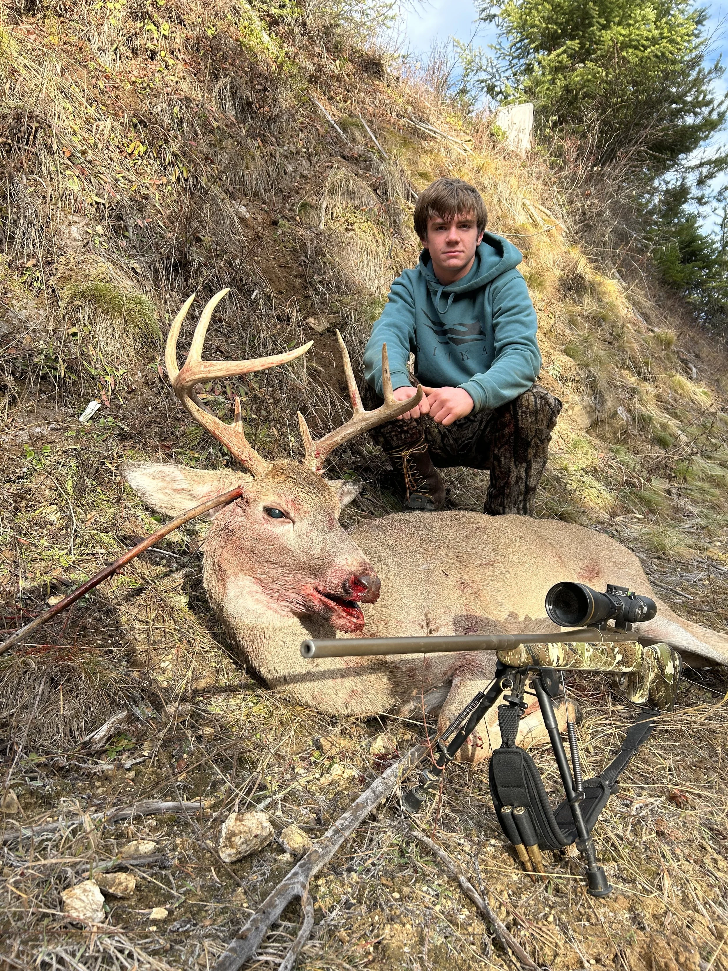 A young boy kneeling next to a large dead deer with antlers, lying on a sloped hillside with grass and rocks. The boy is holding one of the deer's antlers and has a rifle with a scope resting on the ground nearby.
