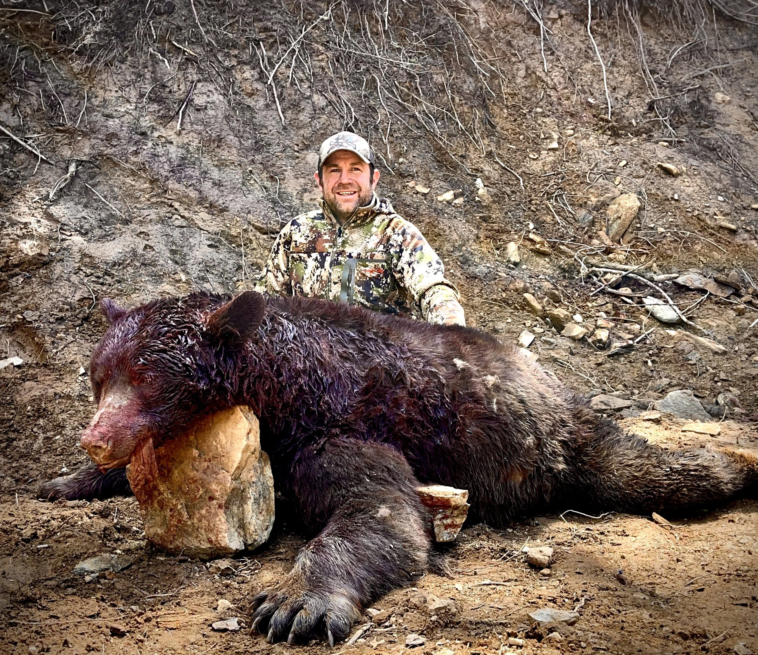 A man in camouflage clothing kneels behind a large, dead bear lying on the ground with a rock under its head. The background is a dirt and rocky hillside.