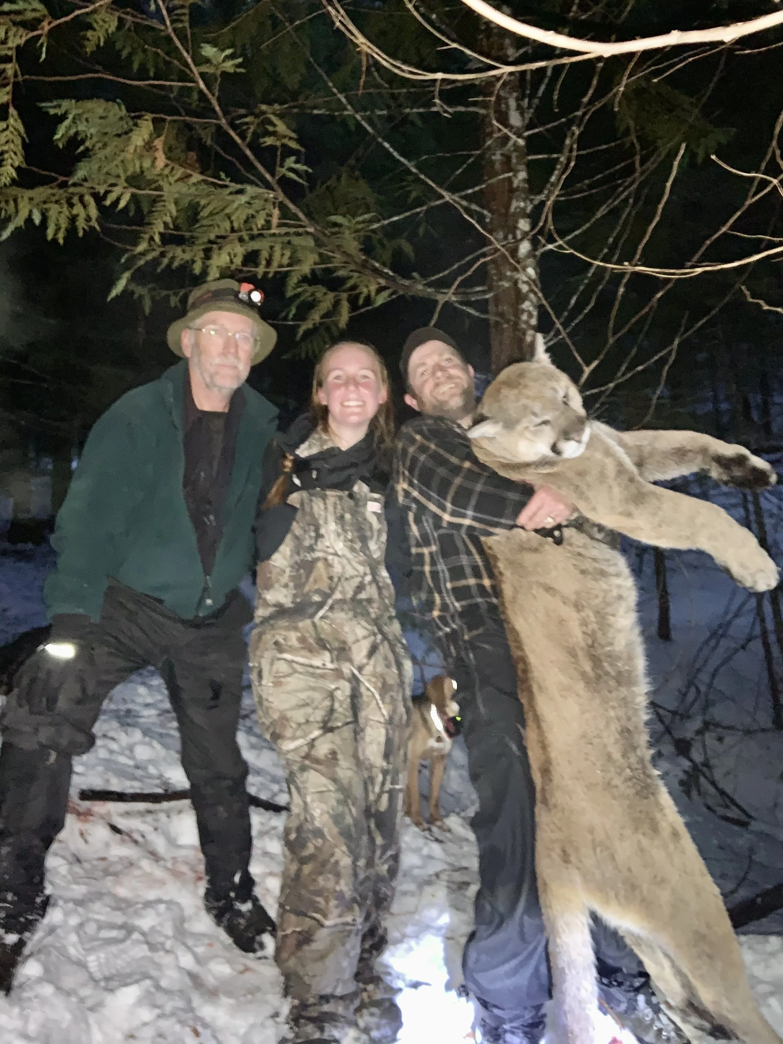 Three people with a lion and a hunting dog in a snowy forest at night, one person holding the lion.