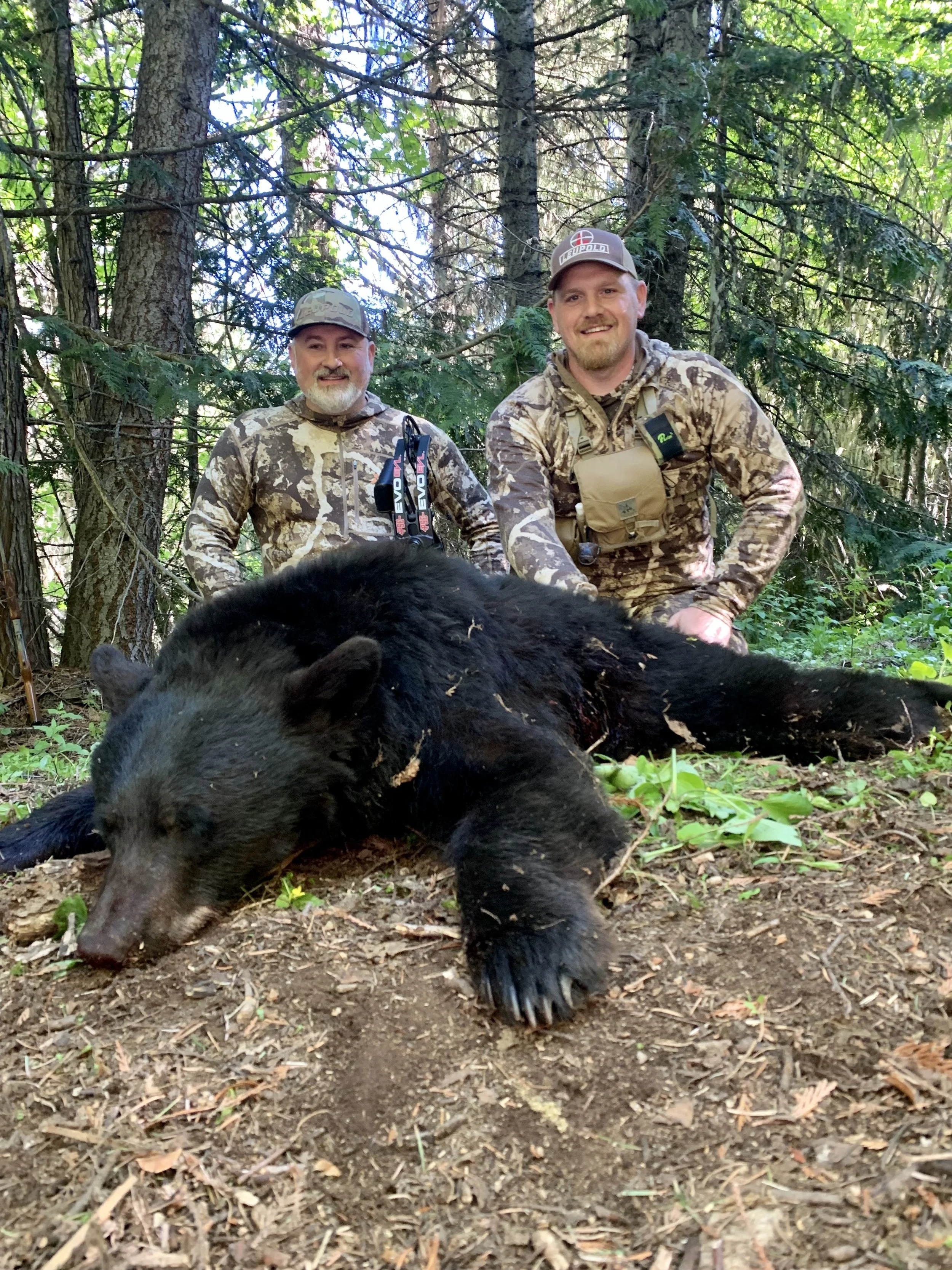Two men in camouflage hunting gear kneel behind a large, black bear lying on the forest floor, with trees and foliage in the background.