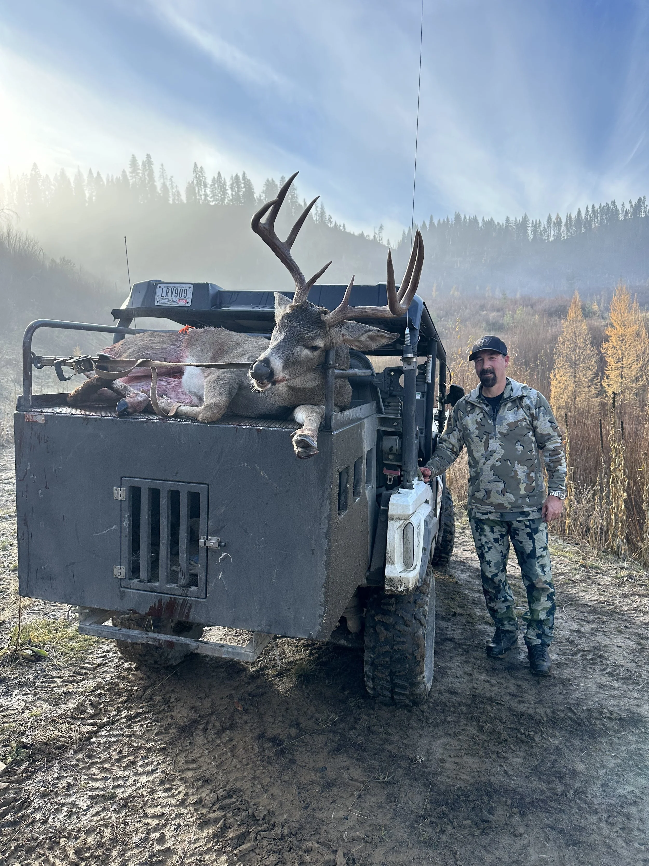 A hunter in camouflage clothing stands next to a utility vehicle with a large mule deer on the back, showing its antlers, in a rural outdoor setting with mountains and trees in the background.