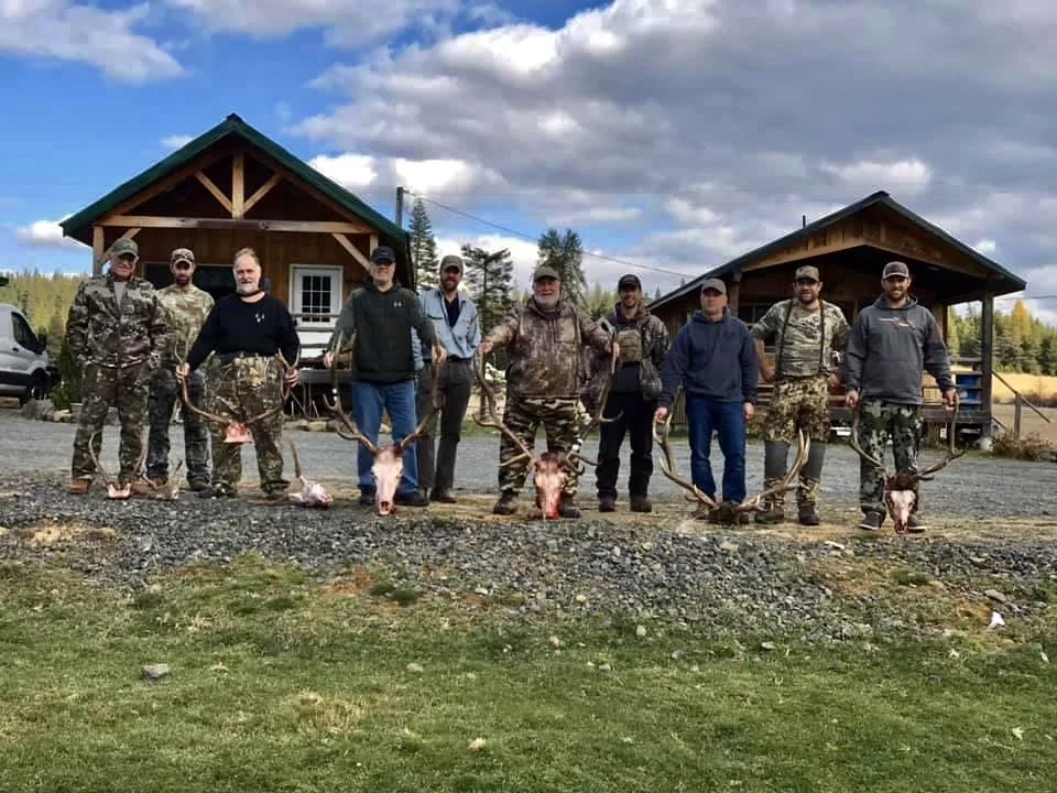 Group of nine men standing outdoors, holding antlers with carcasses attached, in front of two wooden cabins, under a partly cloudy sky.