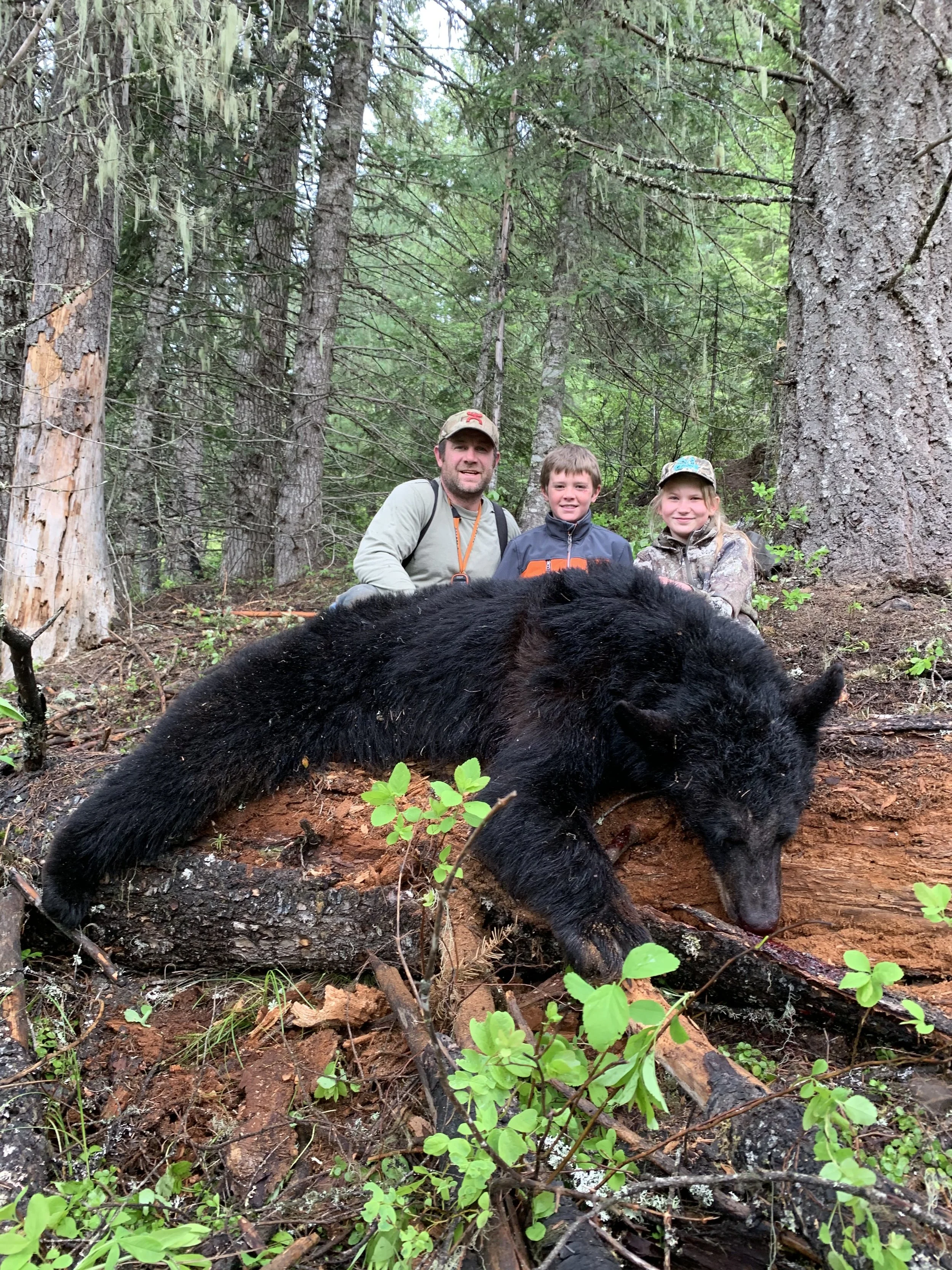Three people, a man and two children, pose behind a large, dead black bear lying on a fallen tree in a forest with green trees and bushes.