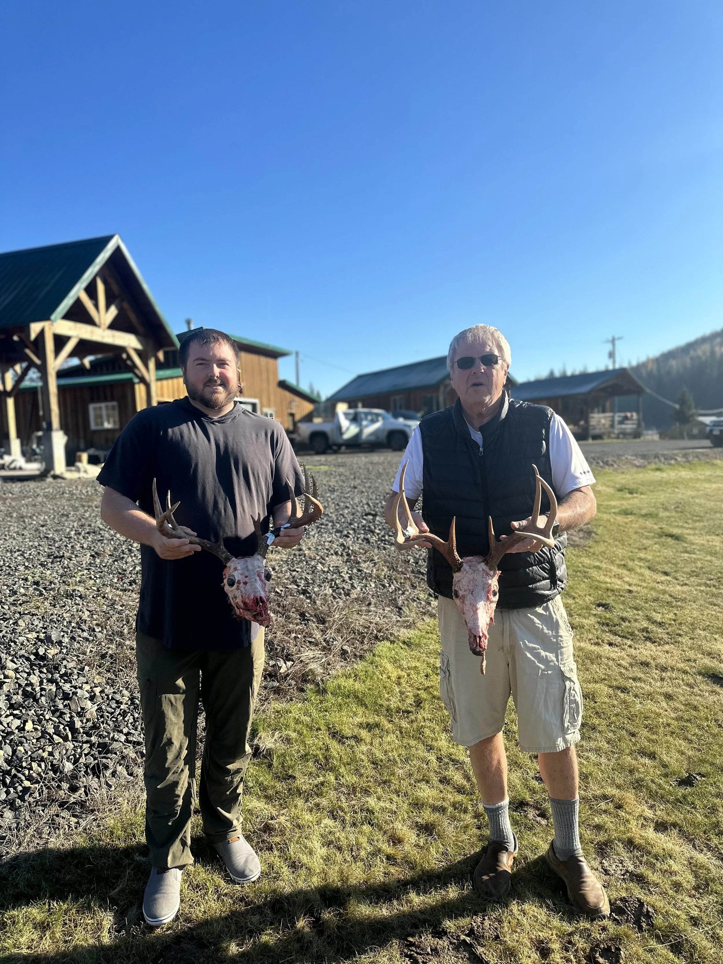 Two men standing outdoors in front of wooden buildings and a gravel area, each holding a butchered deer skull with antlers.