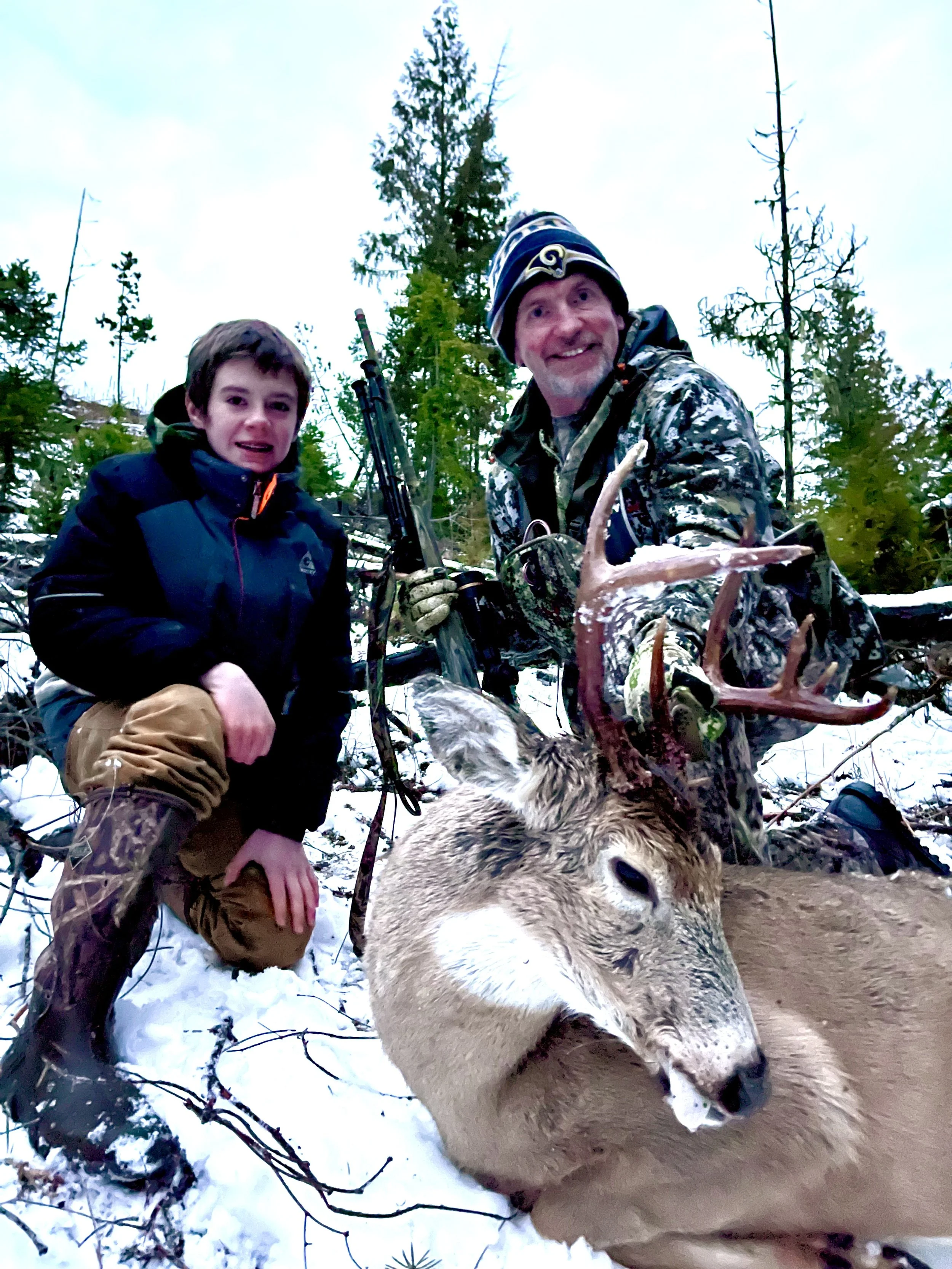 A man and a boy outdoors in a snowy forest, kneeling next to a large deer with antlers. The man is holding the deer's antlers and both are smiling at the camera. The man is wearing camouflage clothes and a beanie, and the boy is dressed in winter gea