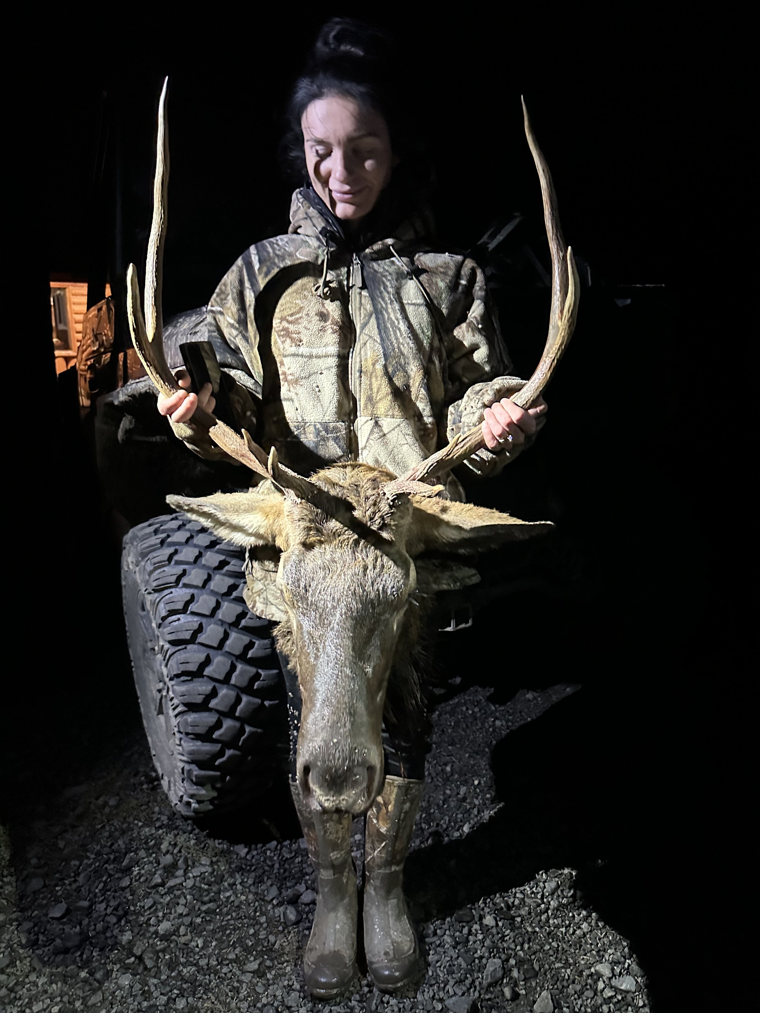Woman in camouflage jacket sitting on a vehicle at night, holding a large moose head with antlers.