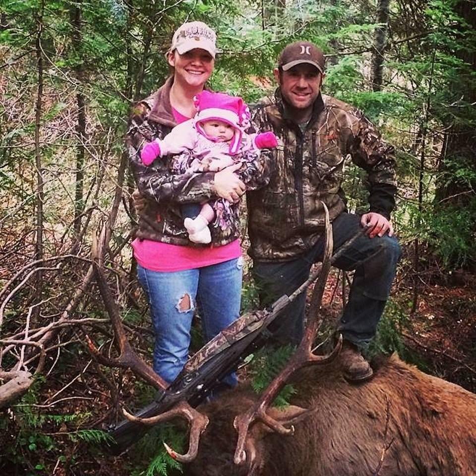 A family of three in camouflage clothing in the woods, posing with a large dead elk that has impressive antlers. The woman is holding a baby girl, and the man is kneeling beside the elk.