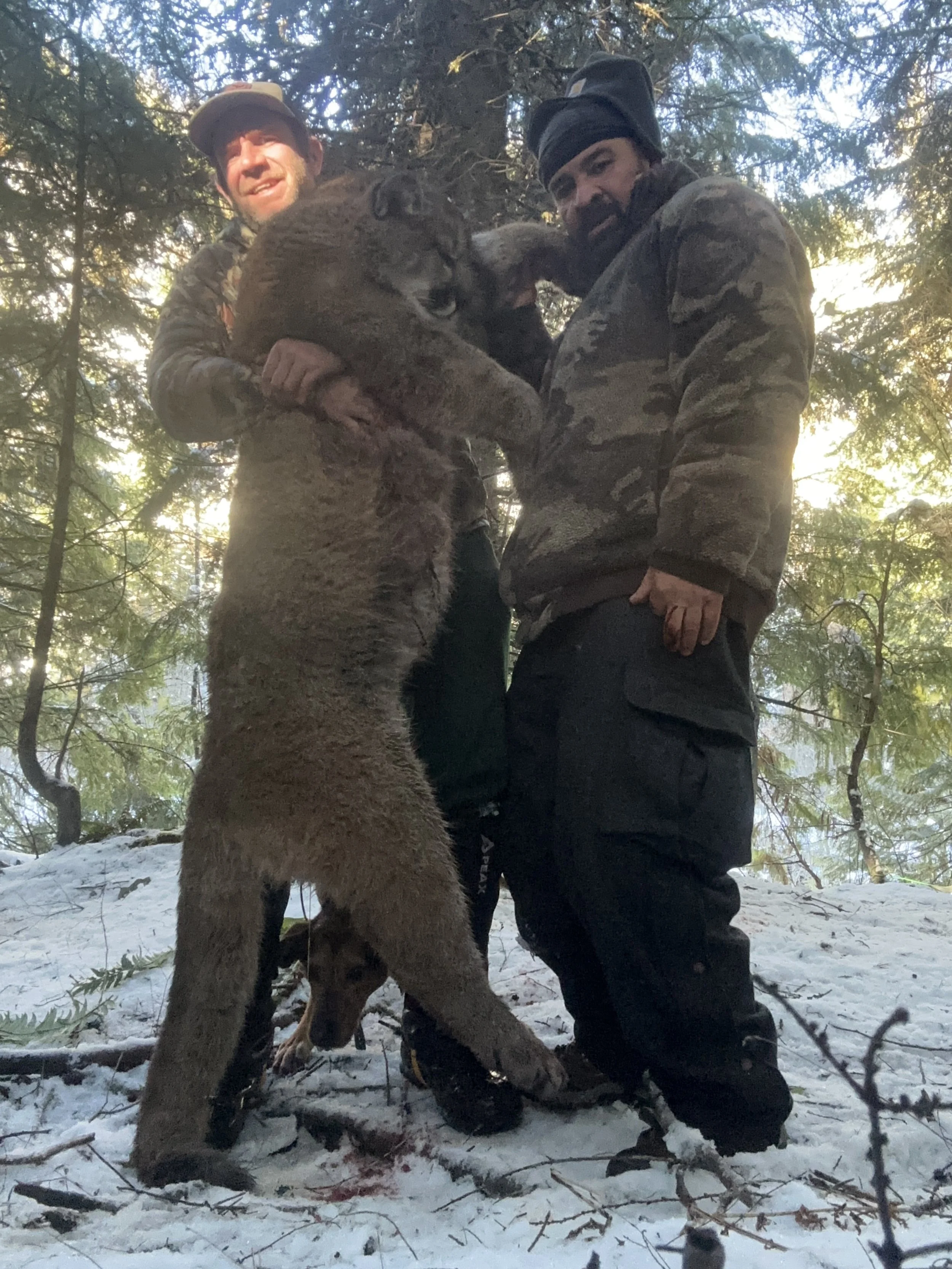 Two men holding a large bear cub in a forest with snow on the ground.