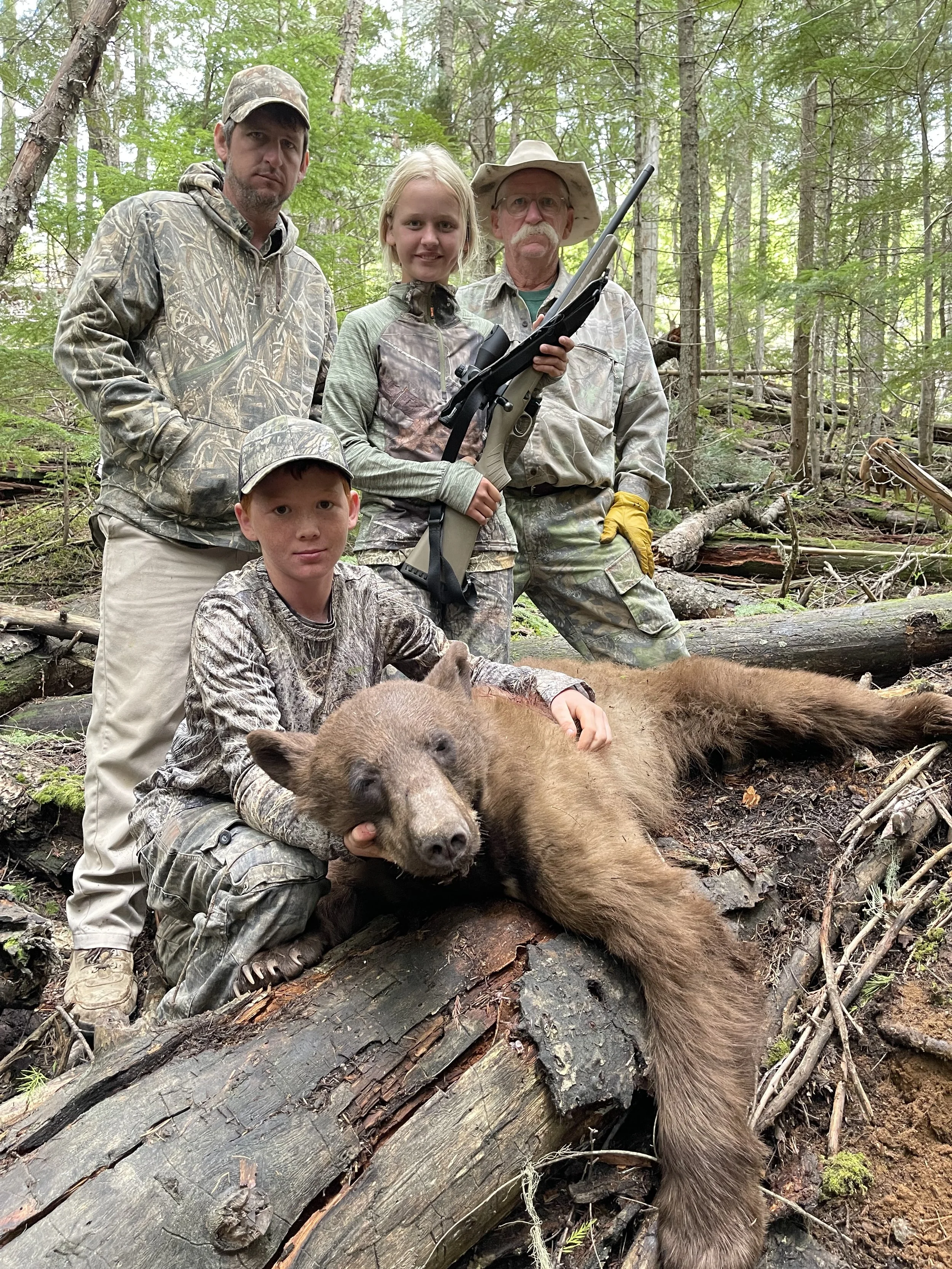 A family of four in camouflage clothing poses in a forest with a dead bear. The father and an elderly man stand behind, the mother and a young boy are seated in front, holding the bear's head and body. The family appears proud of their hunting succes
