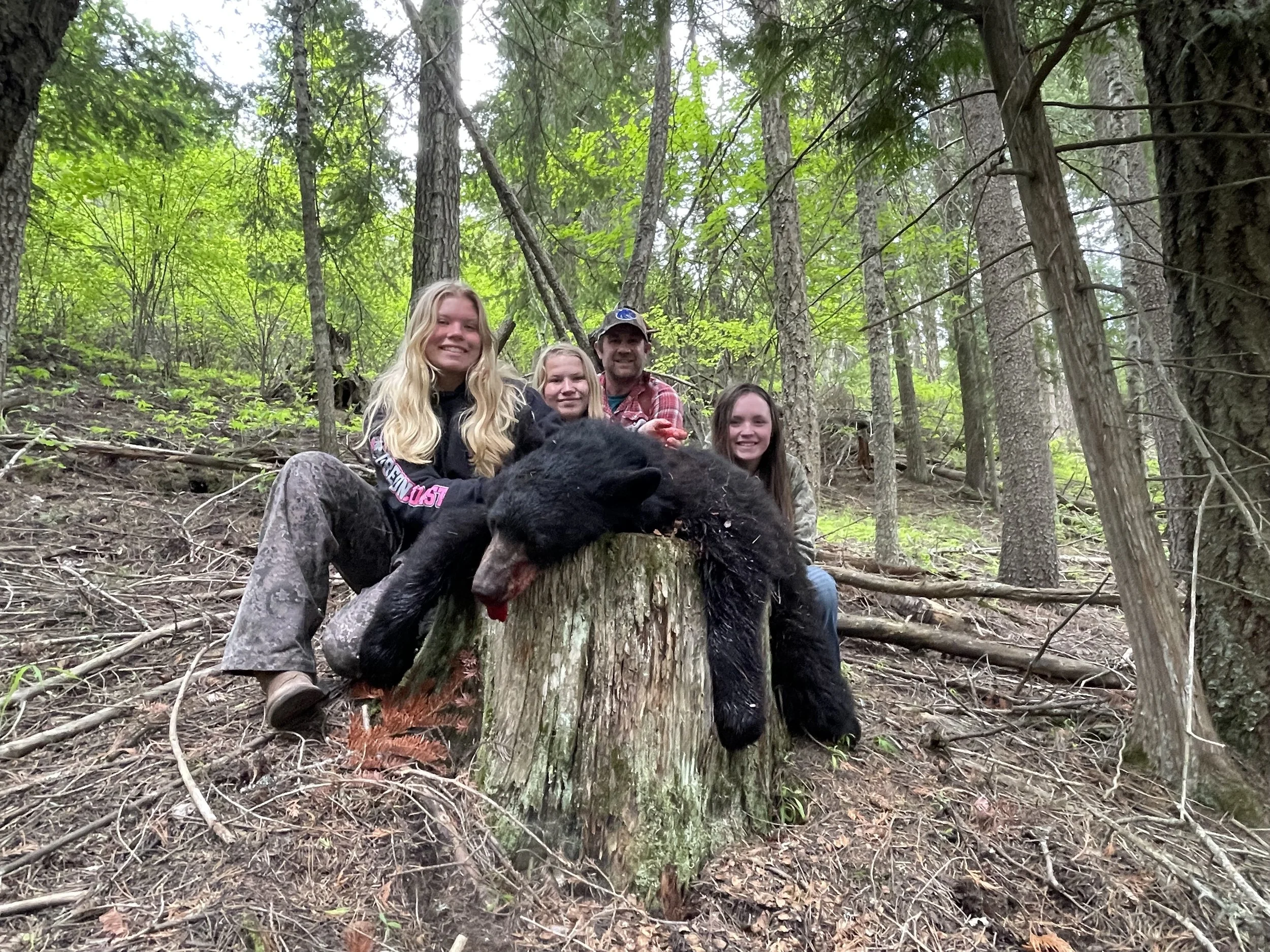 Group of four people, three young women and one man, sitting on a tree stump in a forest, smiling. A black bear cub is draped over the stump with its tongue hanging out.