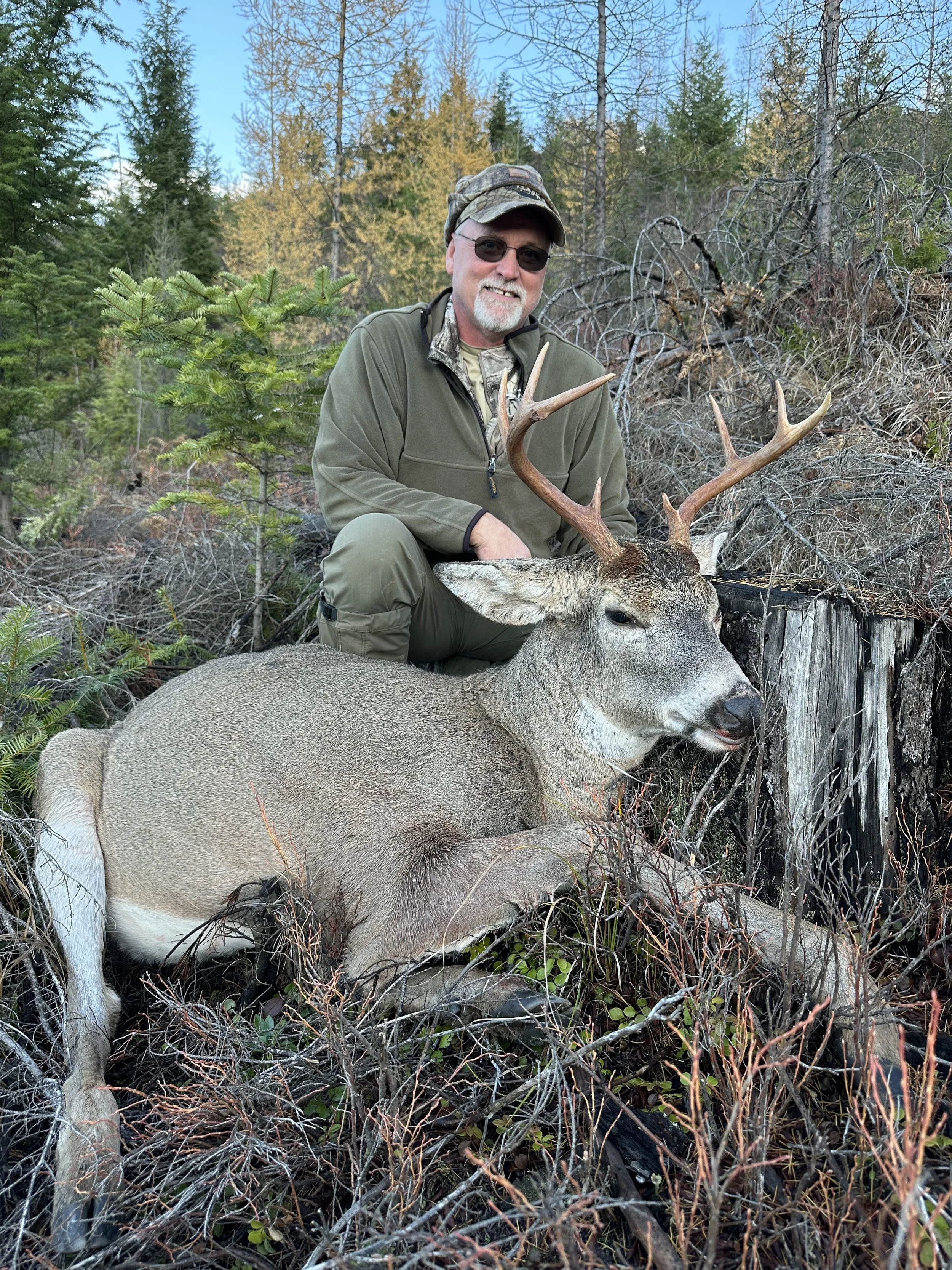 A man kneeling behind a large buck with antlers in a forested area, smiling at the camera.