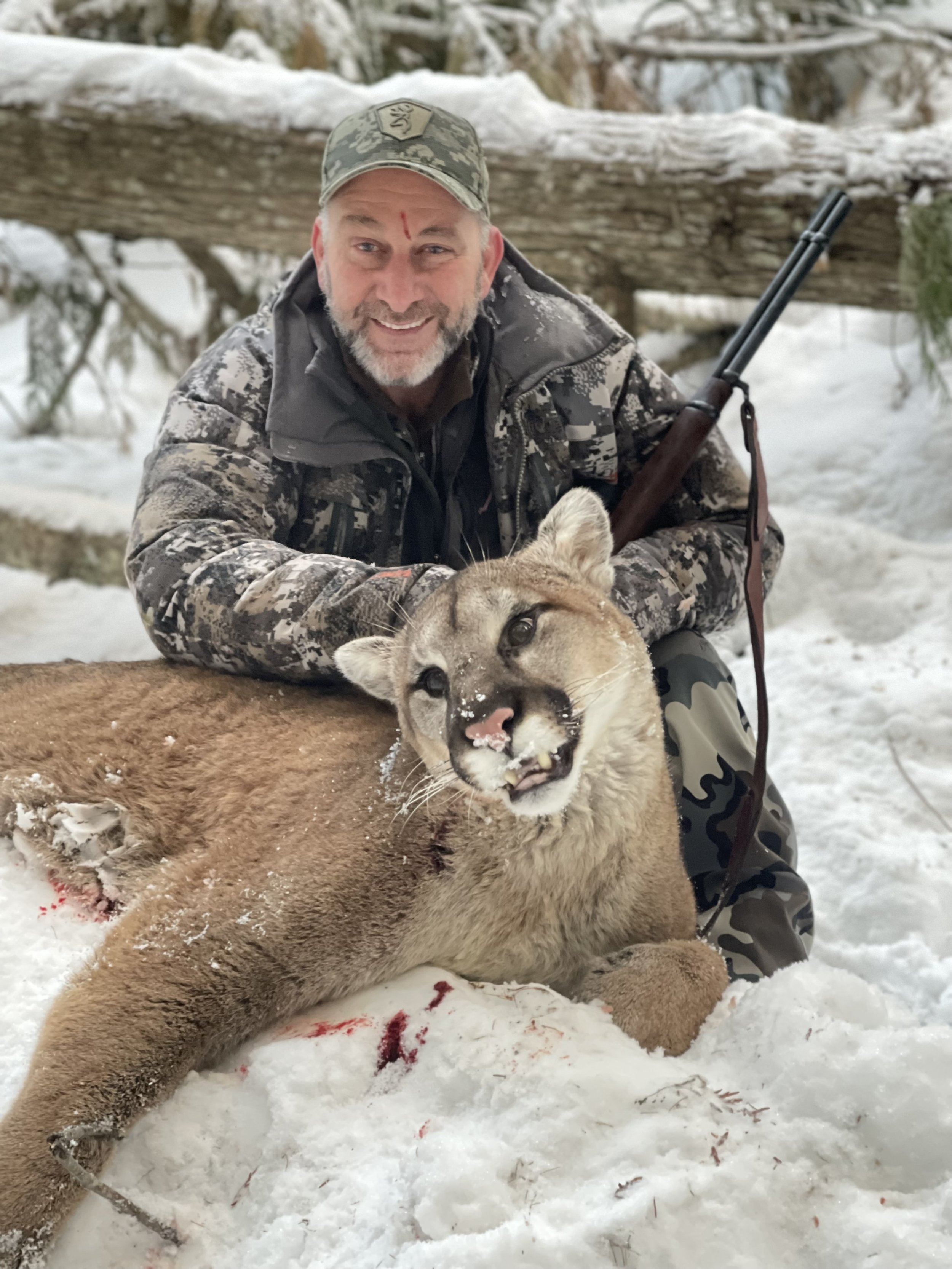 A man in camouflage clothing is smiling while kneeling in the snow next to a dead mountain lion, which has some blood and snow on its face.