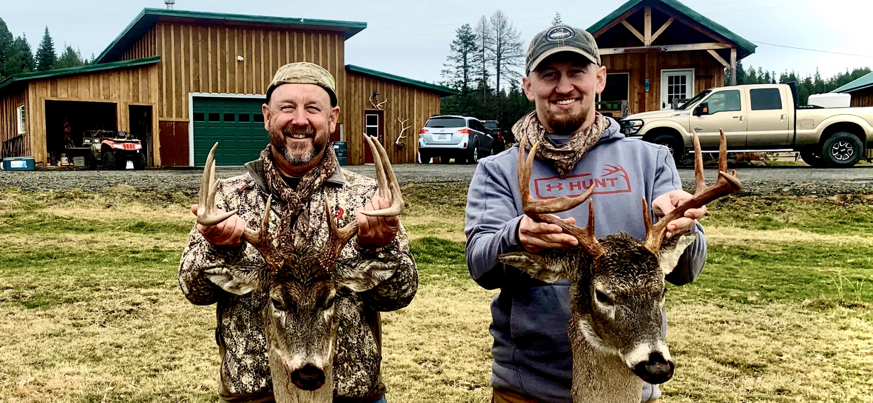 Two men smiling and holding their hunted deer with antlers in front of a wooden house and garage with cars, in a rural outdoor setting.