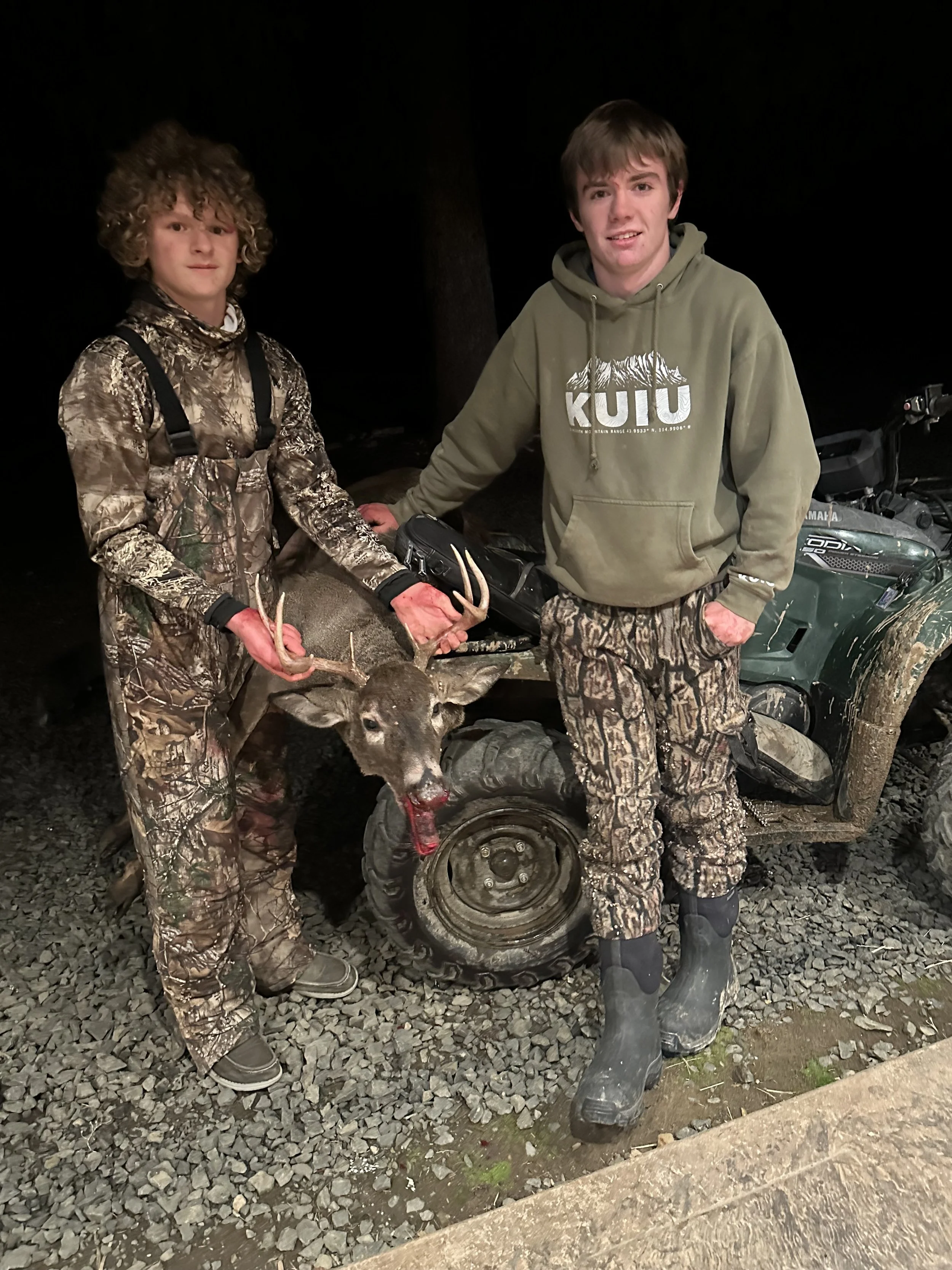 Two young men dressed in camouflage and outdoor clothing standing outdoors at night, proudly holding a large deer they hunted, with the deer’s head resting on the ground. They are standing by an all-terrain vehicle.