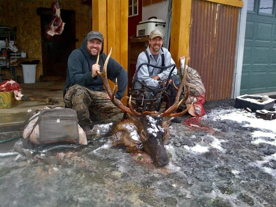 Two men in outdoor attire kneeling next to a large moose with impressive antlers, lying on the ground in front of a wooden building. The scene appears to be outdoors with a muddy, icy ground and some snow visible.