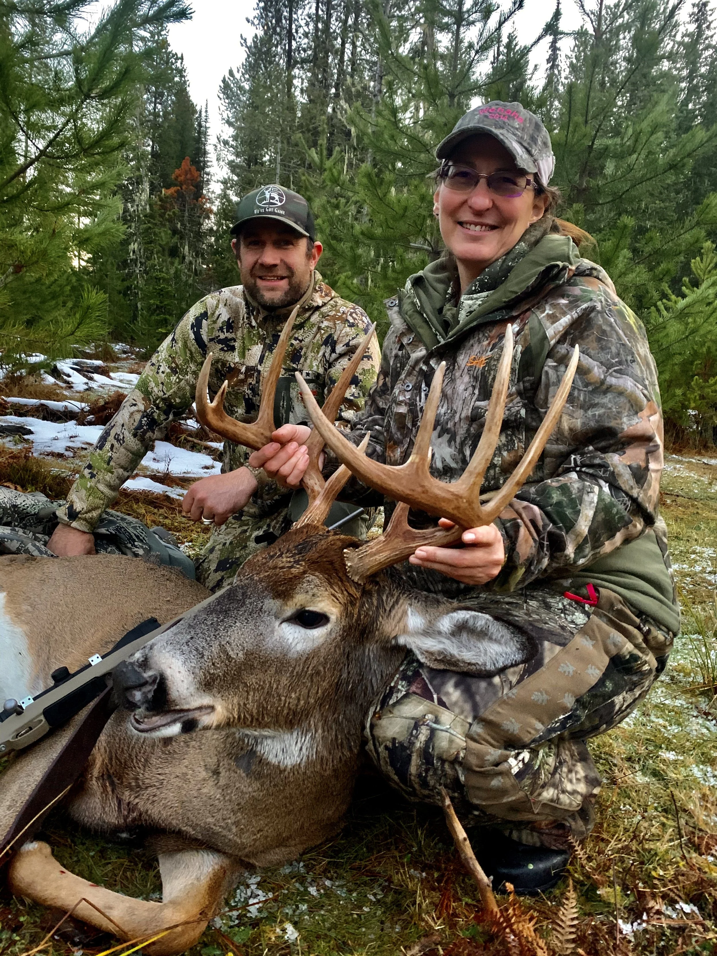 Two hunters, one man and one woman, outdoors in a forest with pine trees, posing with a harvested deer that has large antlers, both smiling at the camera.