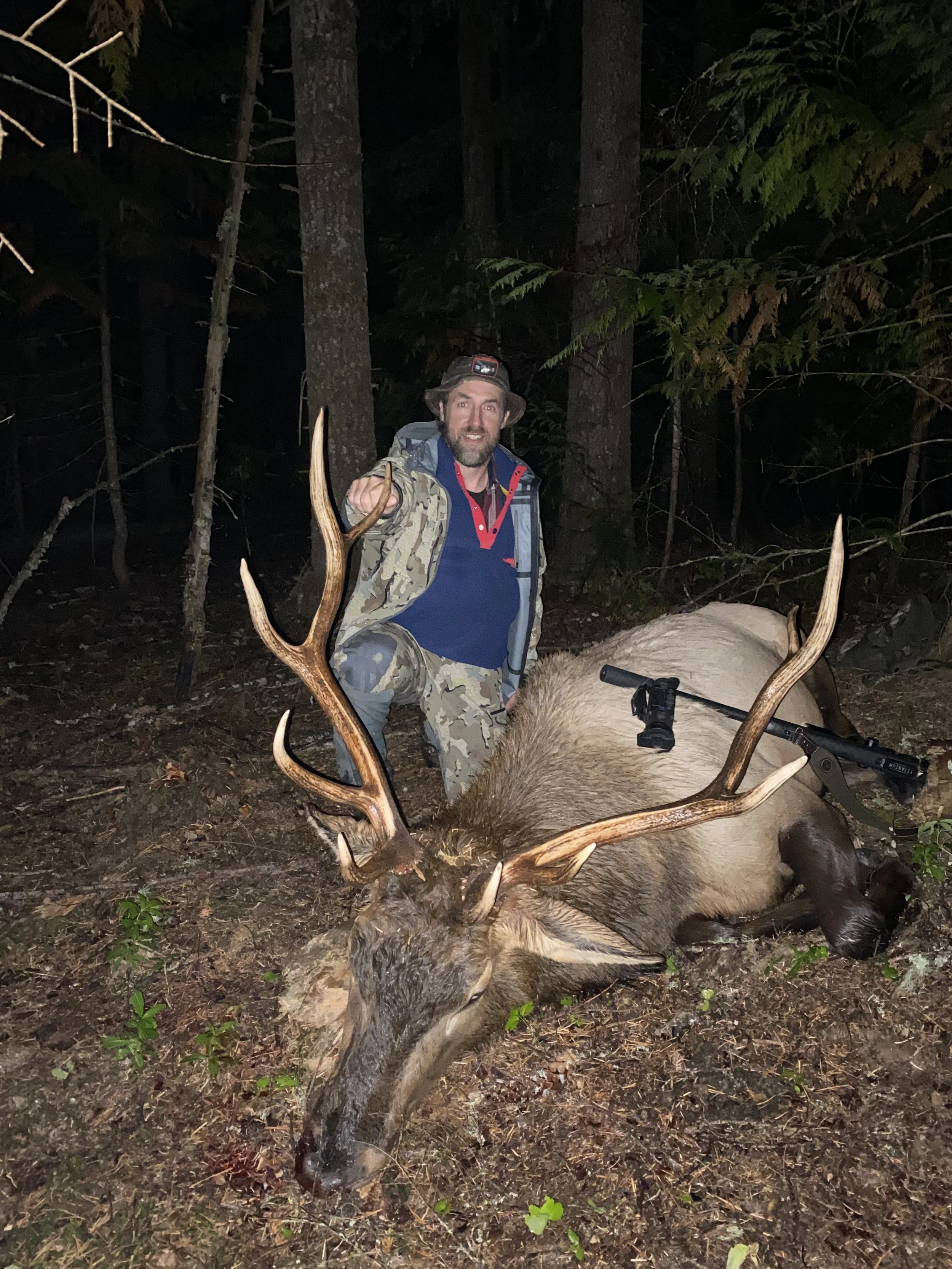 A man in camouflage clothing and a hat kneeling next to a large elk he has hunted in a forest at night, with the elk lying on the ground.