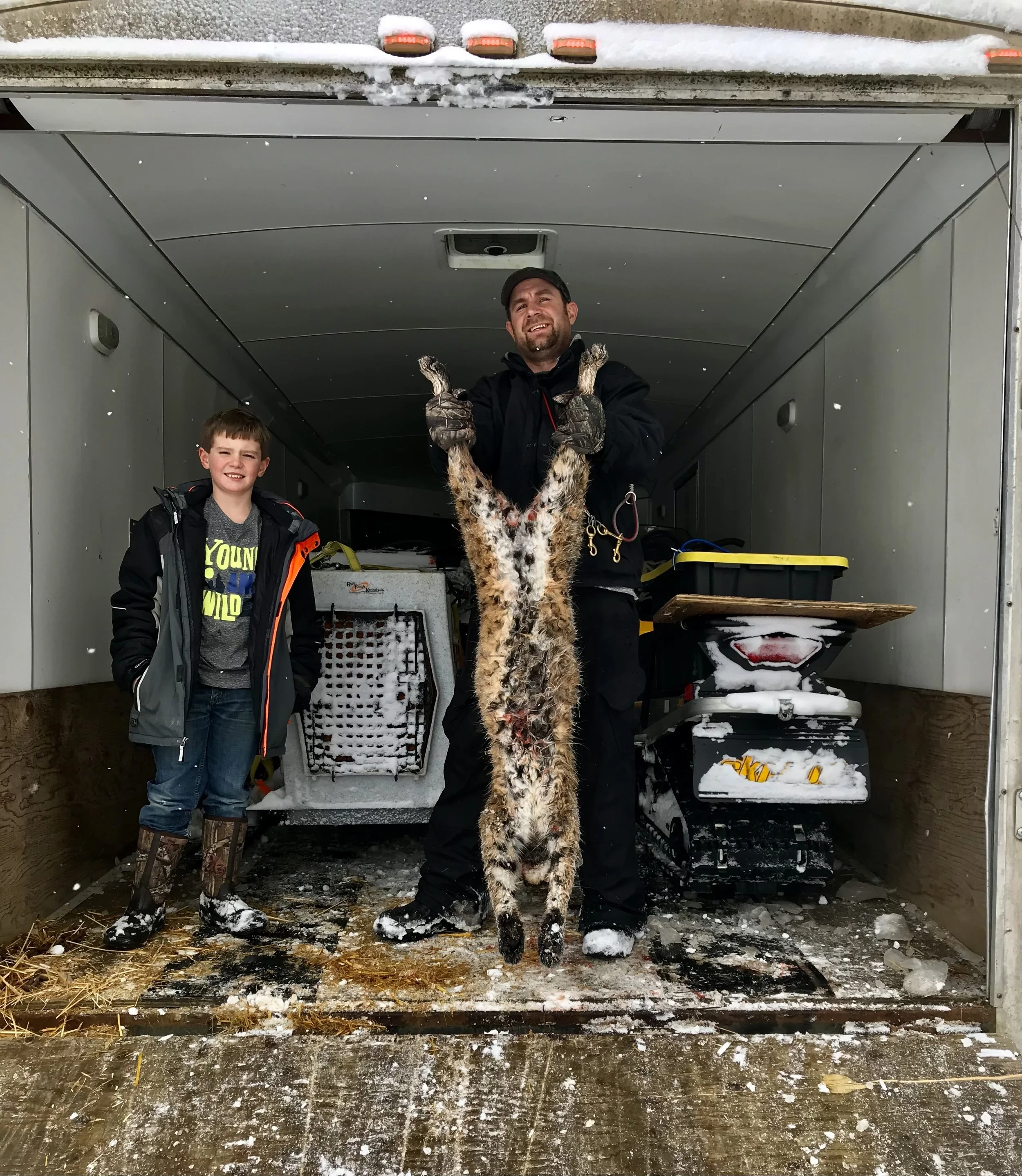 Two boys and a man standing in a snow-covered trailer, holding a bobcat. The man is smiling, and one boy is smiling, wearing winter clothing and boots. Inside the trailer, there is equipment and a small tracked vehicle.