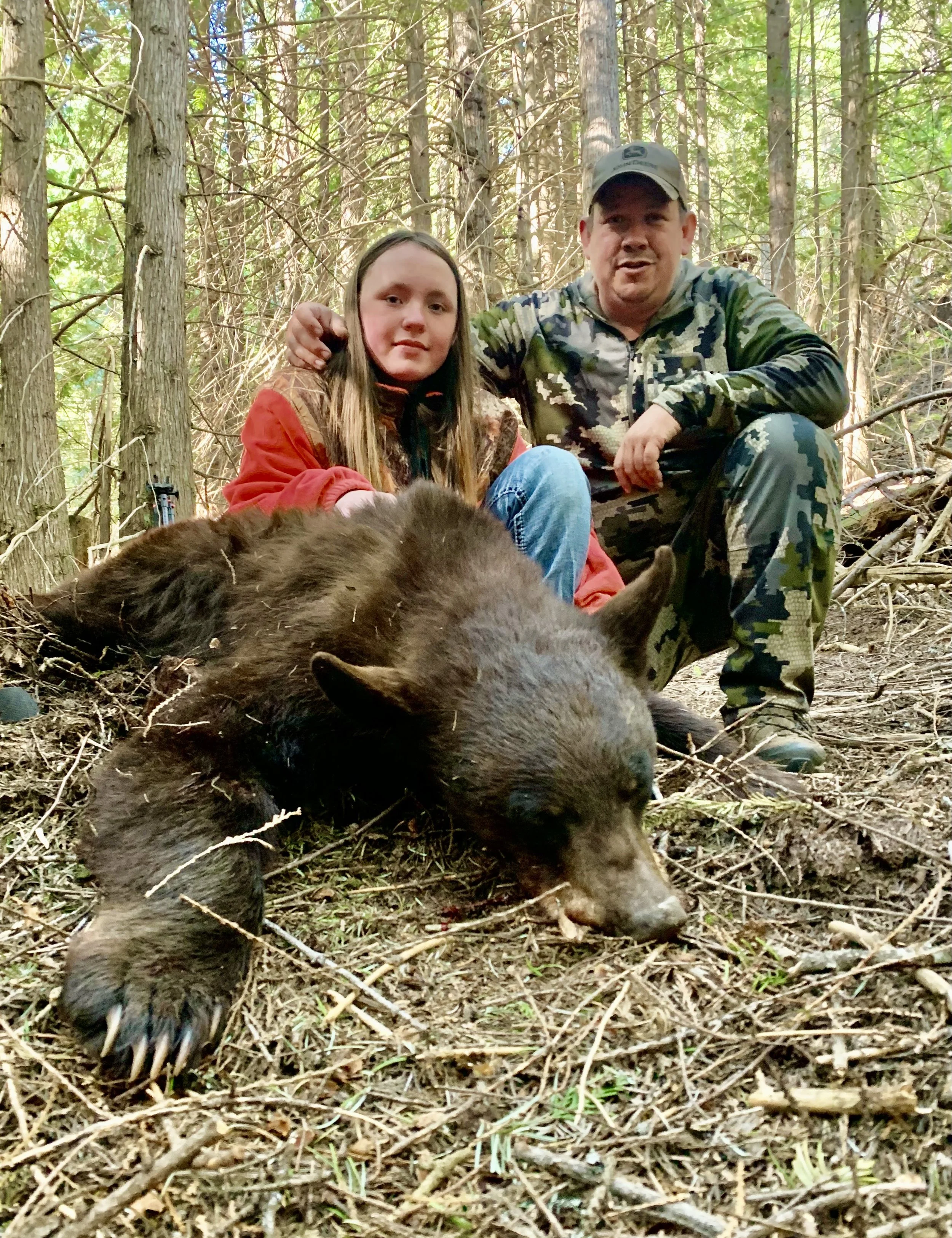 A man and a girl sitting in a forest beside a recently killed black bear.