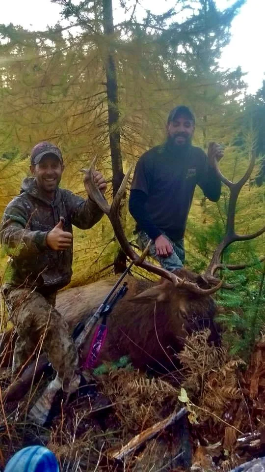 Two hunters posing with a large elk in a forested area, one giving a thumbs-up, both smiling, in autumn surroundings.