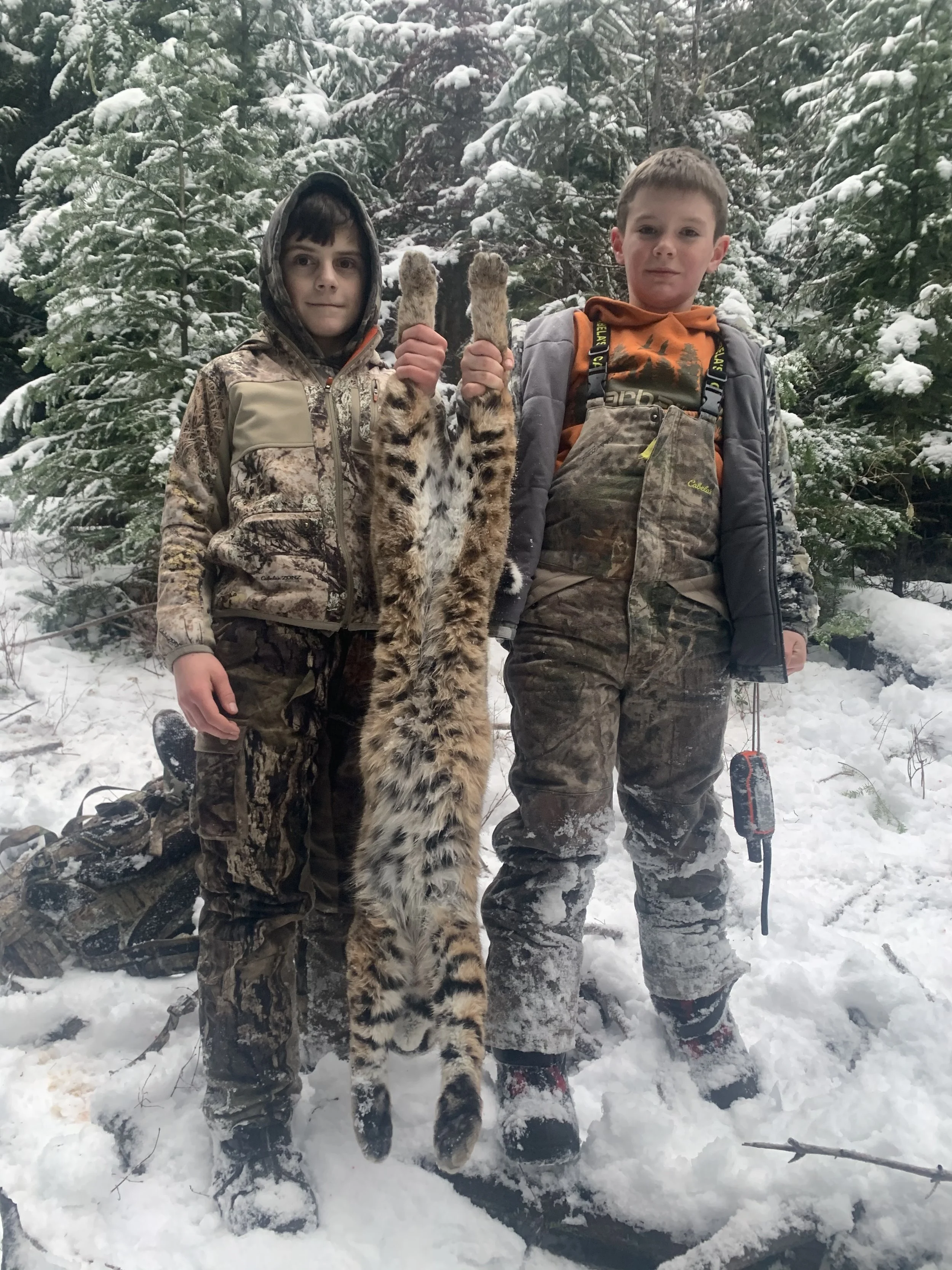 Two young boys in camouflage clothing standing in snow-covered forest, holding up a caught animal, possibly a bobcat, hanging upside down.