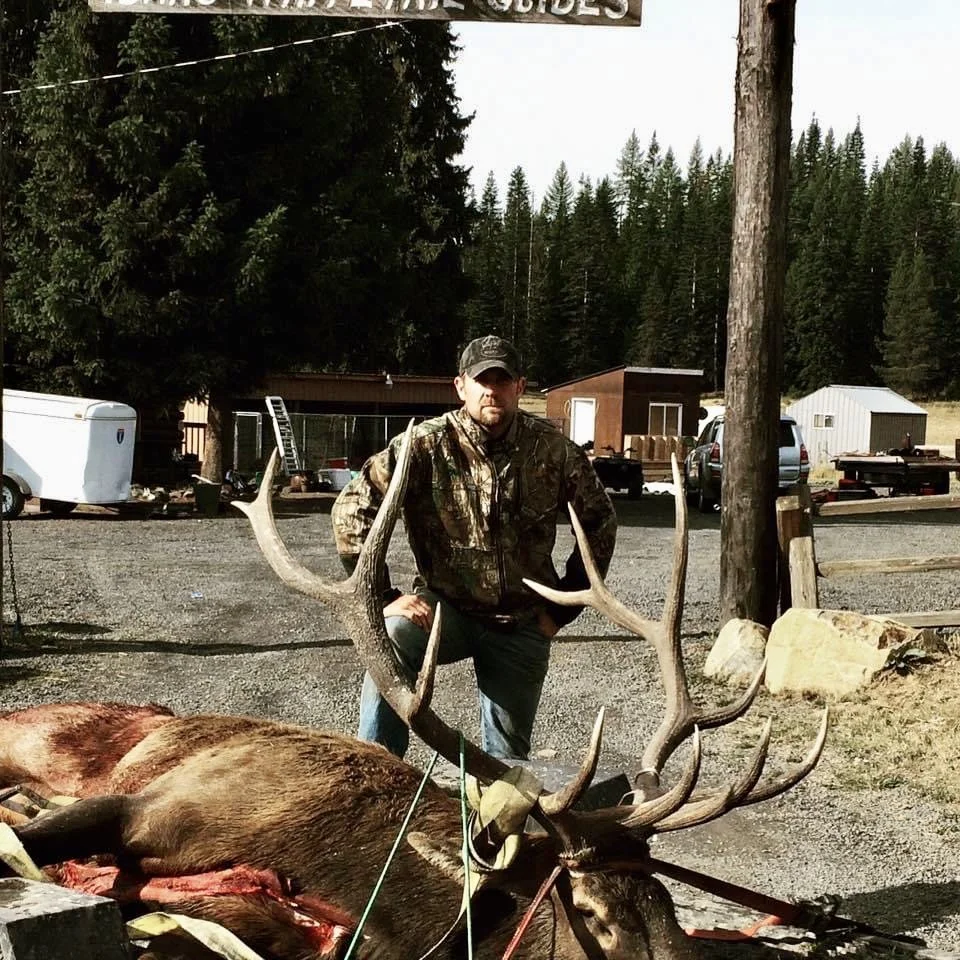 A man in camouflage clothing standing behind a large dead elk with prominent antlers, outdoors in a rural area with trees, buildings, vehicles, and a gravel ground.