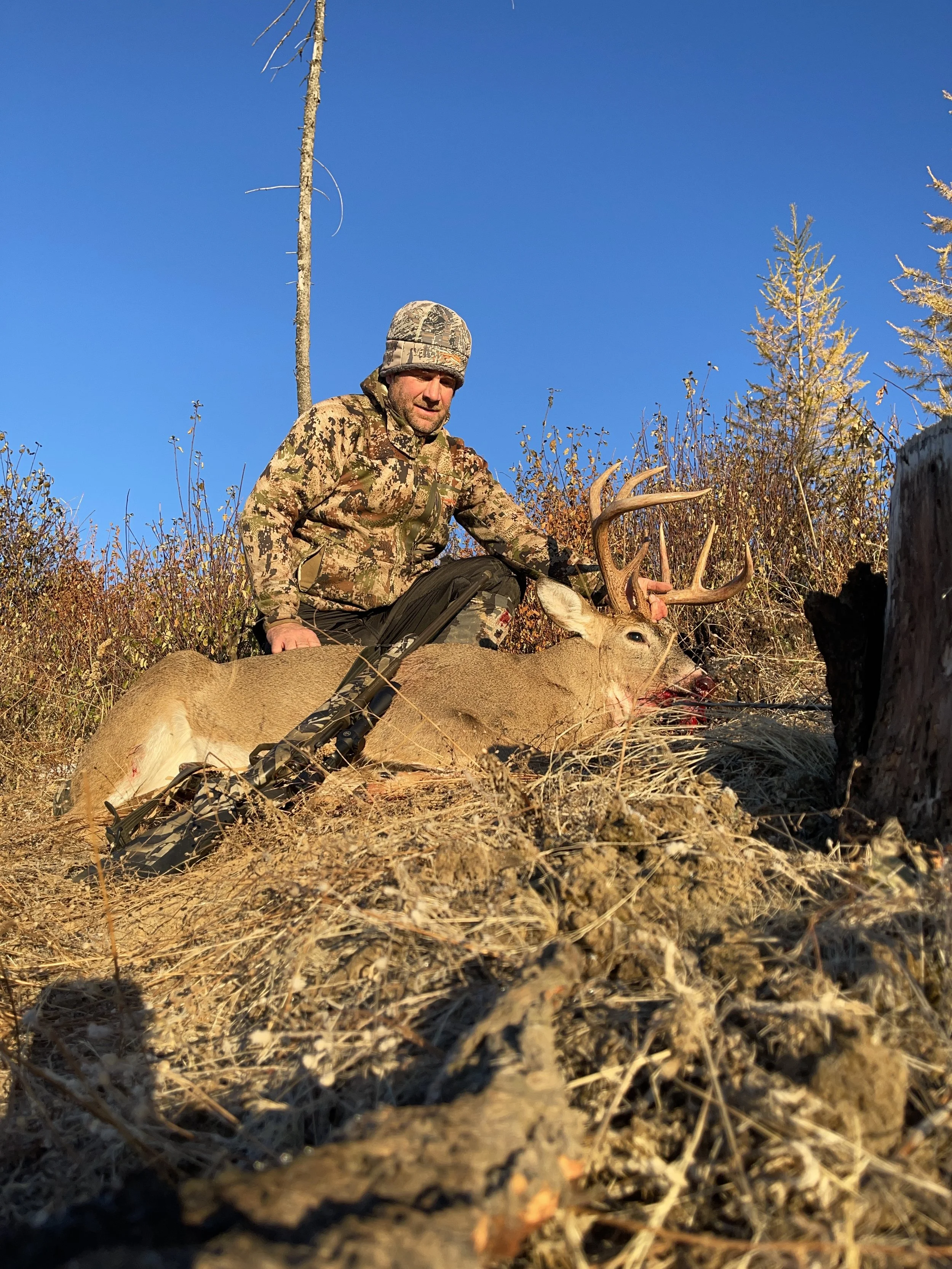 A man dressed in camouflage hunting gear kneels next to a large deer with antlers, which he has just hunted. The man is holding the deer's antlers and appears to be in a natural outdoor setting with dry grass, shrubs, and trees under a clear blue sky