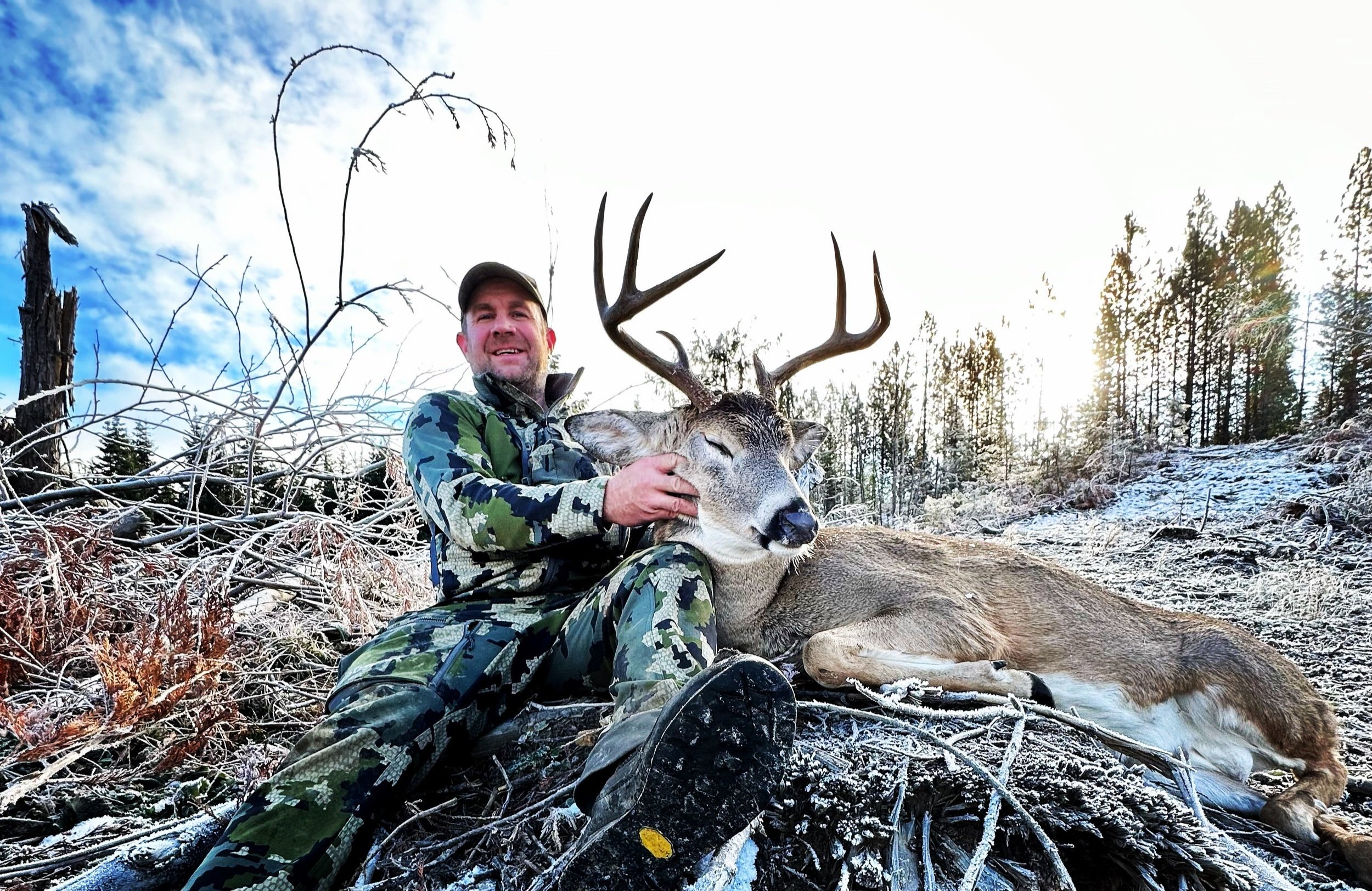 A man in camouflage clothing sitting on snow-covered ground, holding a large deer with antlers. The scene is outdoors in a forest with some trees and a partly cloudy sky.