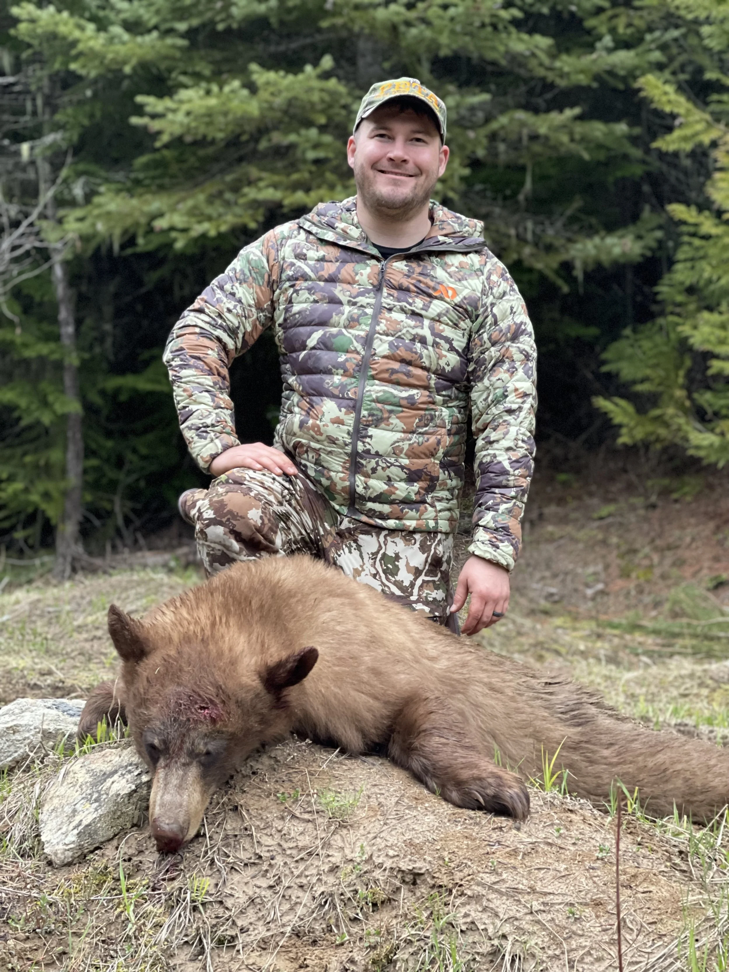 A man in camouflage clothing kneels next to a dead bear lying on the ground outdoors with trees in the background.