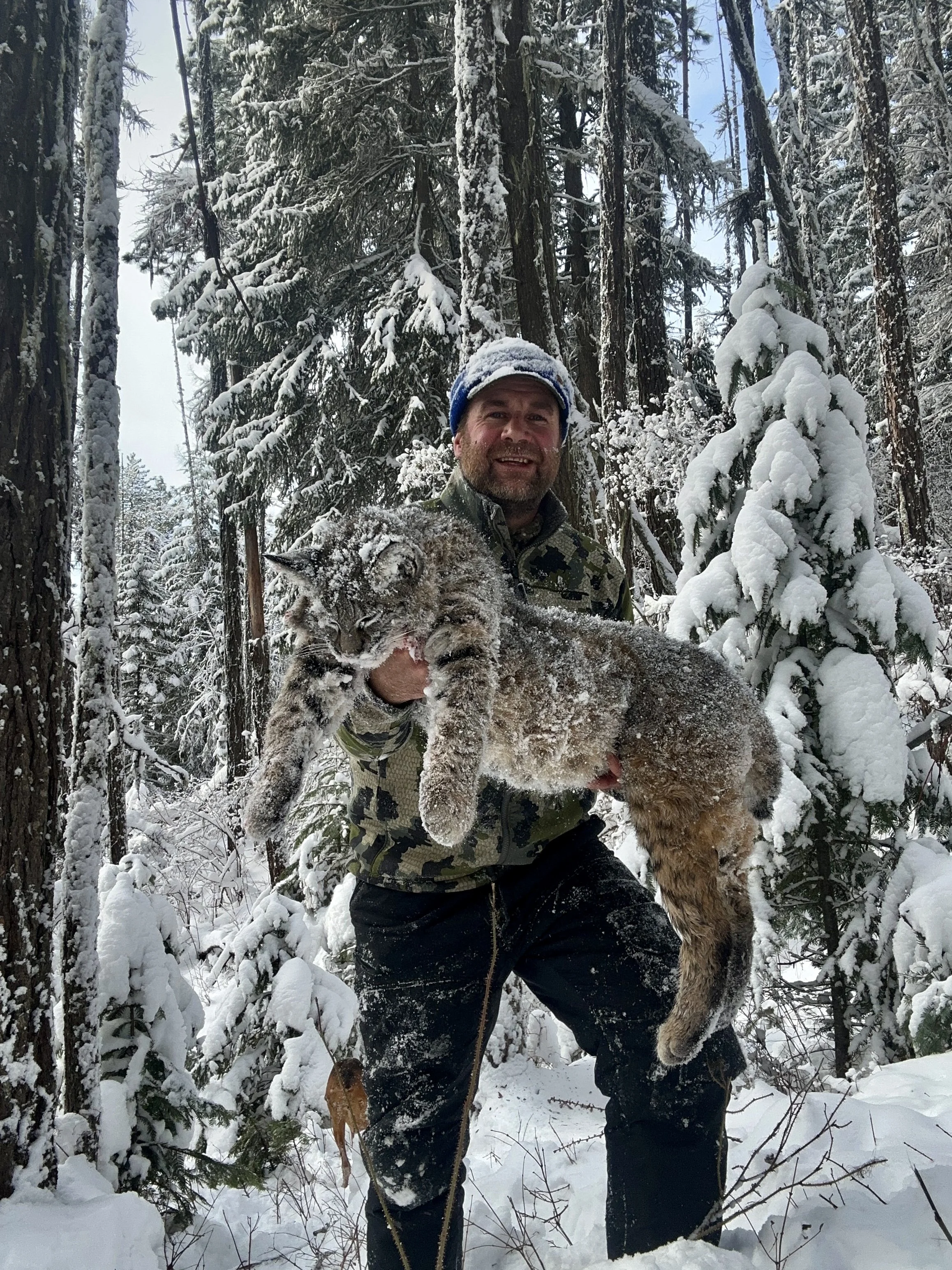 A man in camouflage clothing holding a large, snow-covered mountain lion in a snowy forest with tall trees and snow-covered branches.