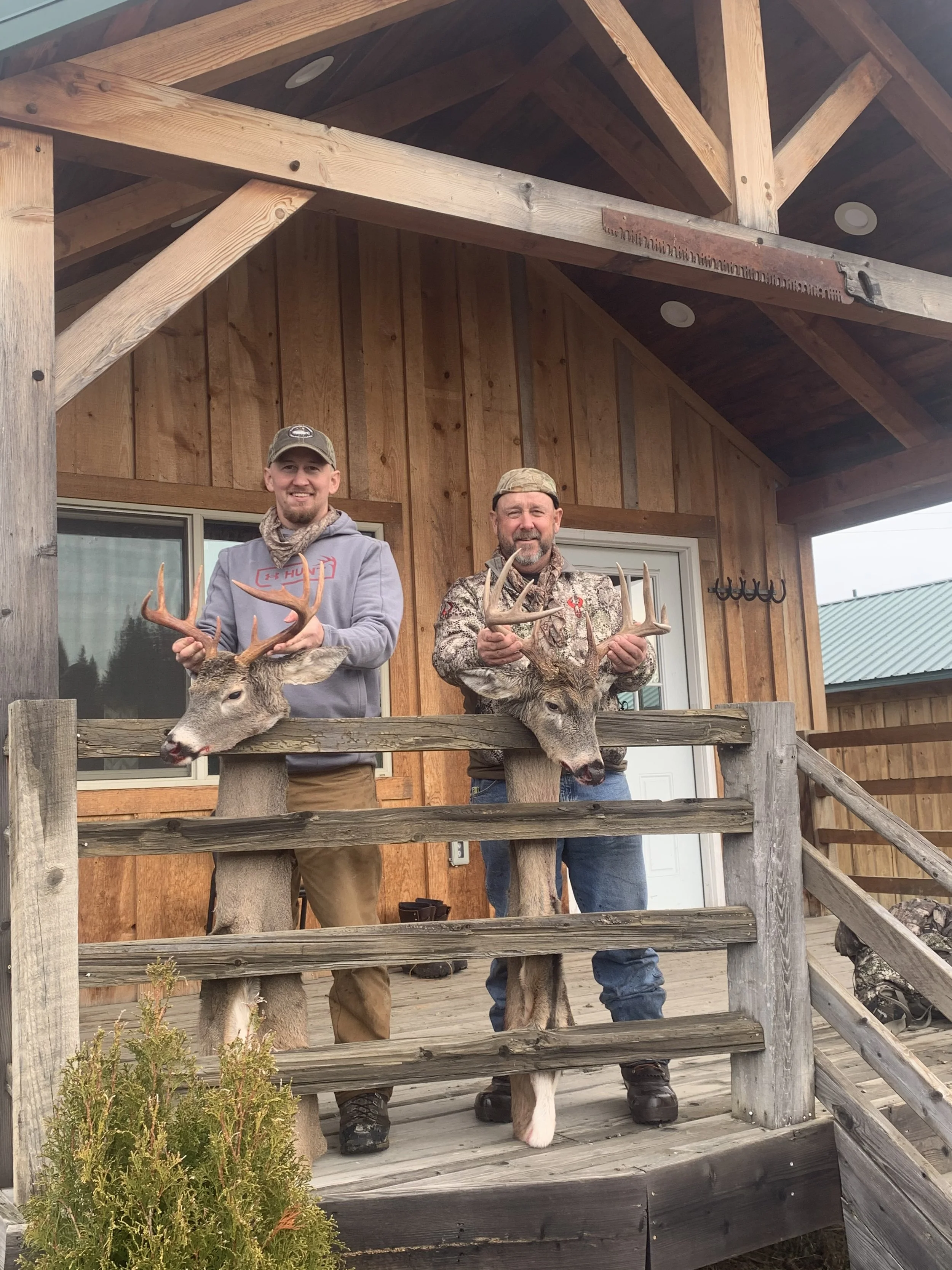Two men standing on a wooden porch holding deer heads with antlers, showcasing their hunting trophies.