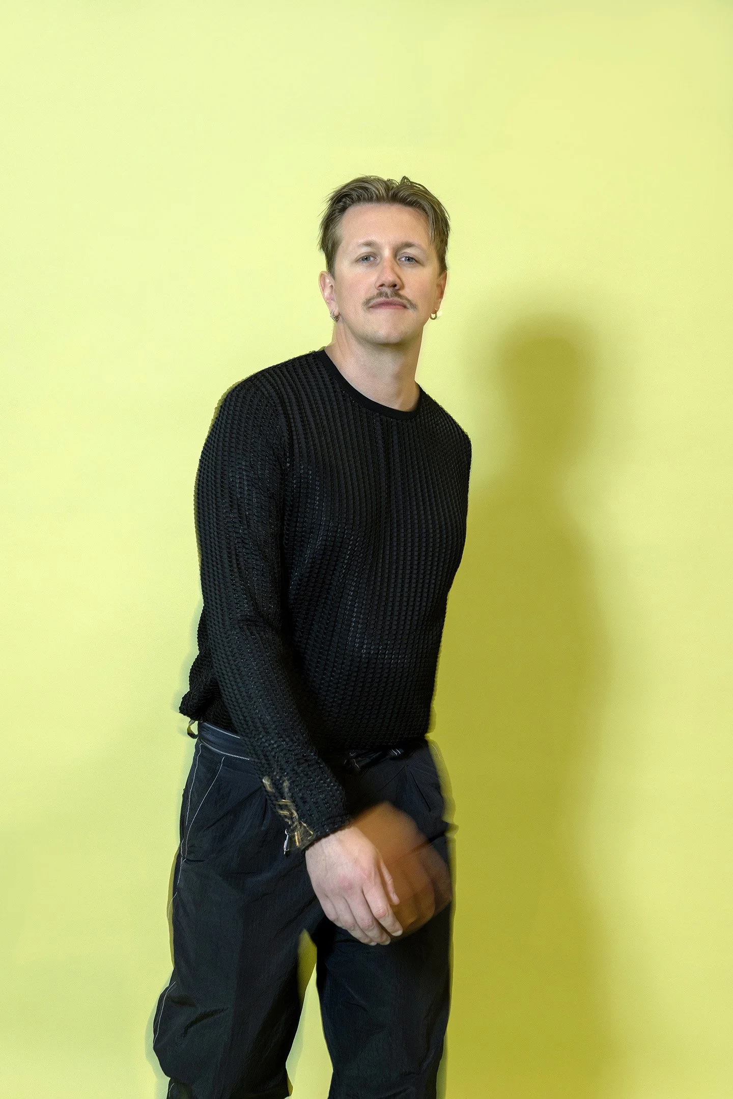 En person med blond hår, kort skæg og mustache, iført sort sweater og sorte bukser, står foran en lys gul baggrund.