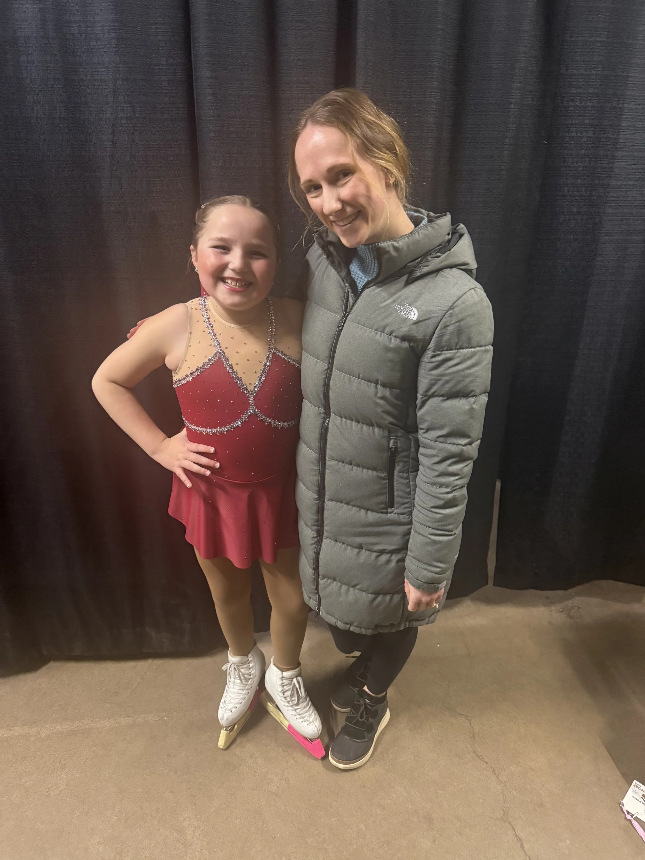 A young girl in a red figure skating dress with rhinestone embellishments, standing on ice skates, next to an adult woman in a gray North Face winter coat, both smiling and posing together in front of a dark curtain.