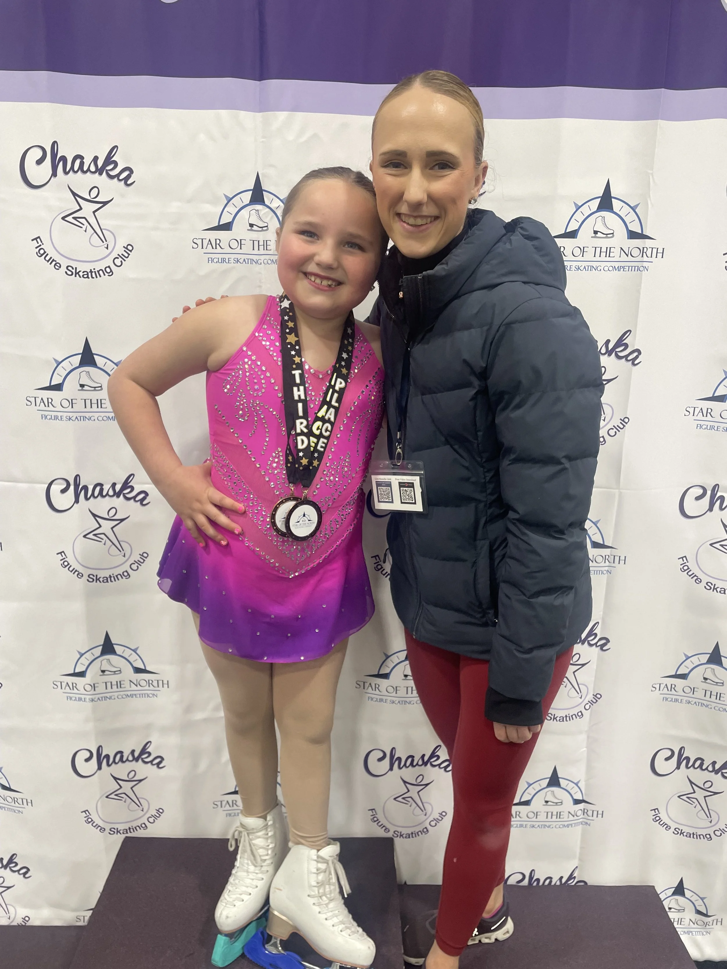 A young girl in a pink figure skating dress with rhinestones and beige tights, wearing a medal, stands next to an adult woman in a dark blue puffer jacket and red leggings. Both are smiling and posing for a photo on a black platform, with a backdrop featuring the logos of Chaska Figure Skating Club and Star of the North Figure Skating Competition.