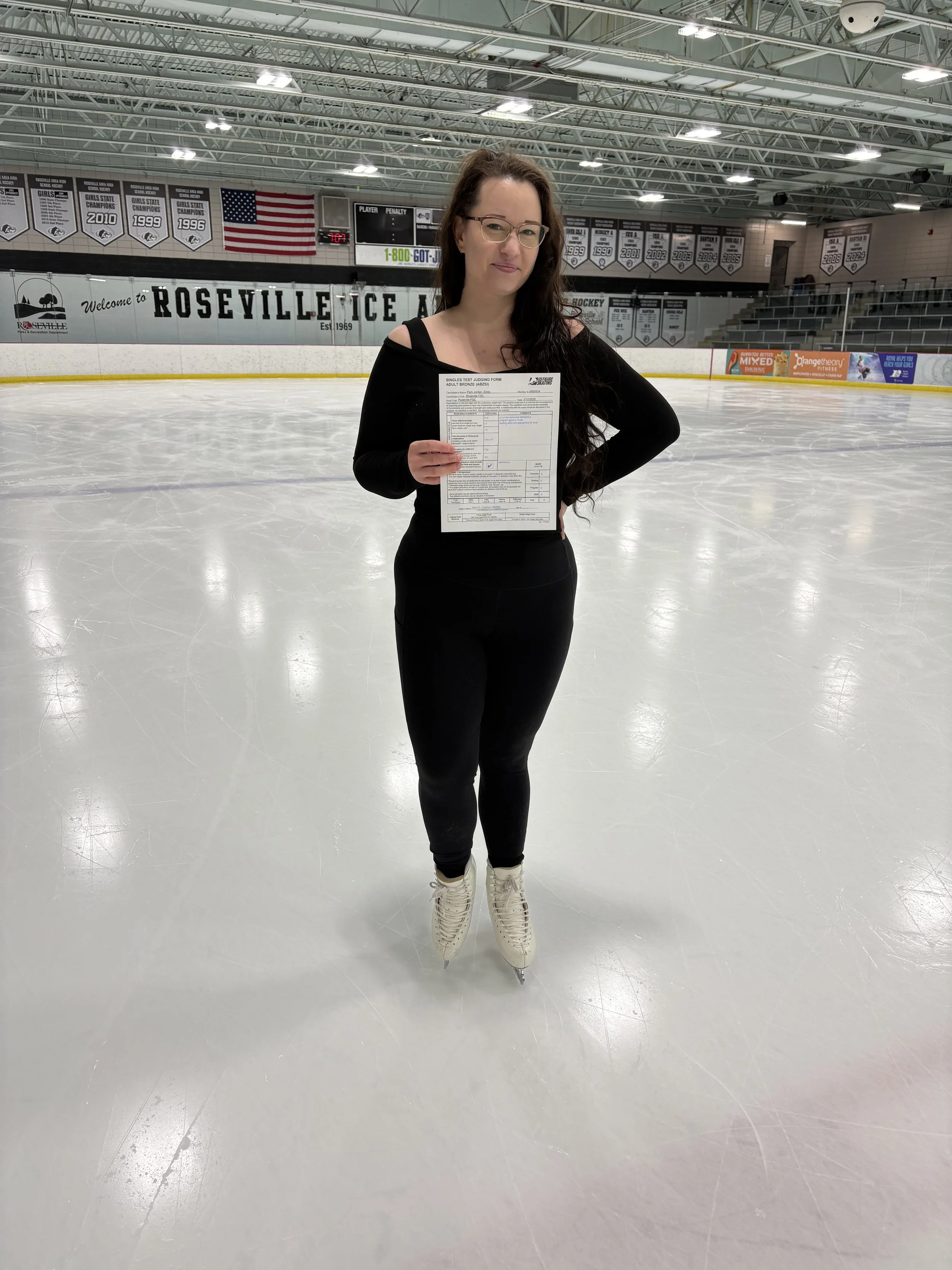 Woman in black skating outfit holding a form at an ice skating rink with banners and flags in the background.