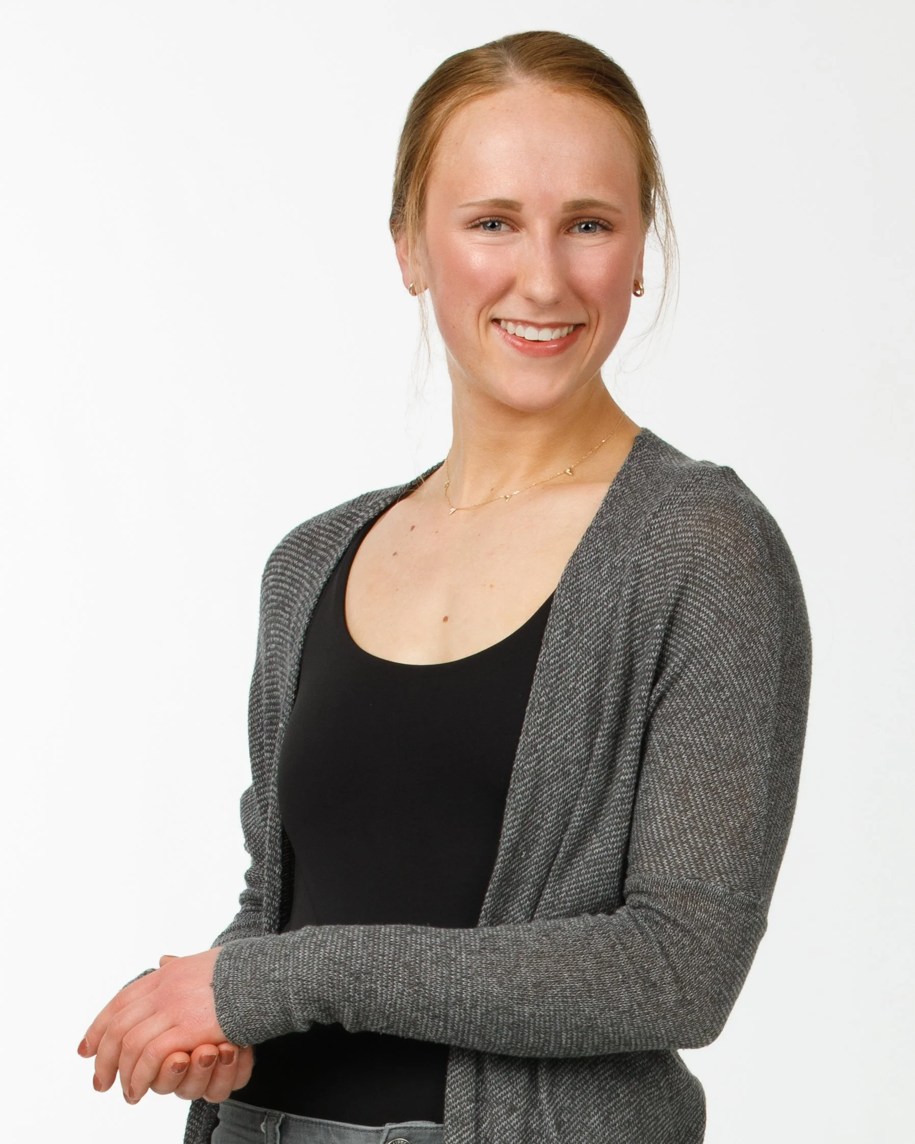 A young woman smiling, with reddish hair tied back, wearing a black top, gray cardigan, and jewelry, standing against a plain white background.