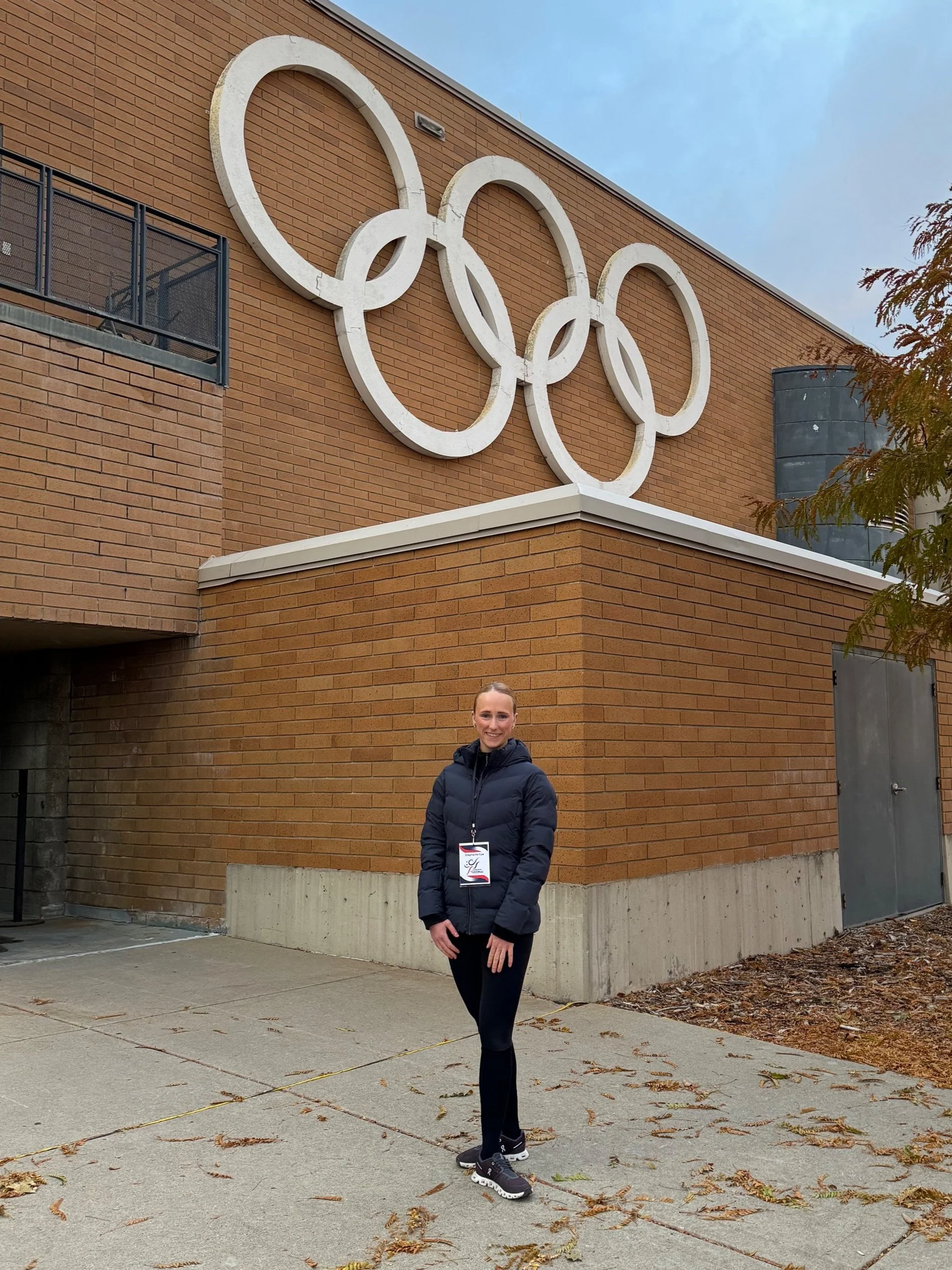 A woman in athletic clothing standing outside near the Olympic rings mounted on a brick building.